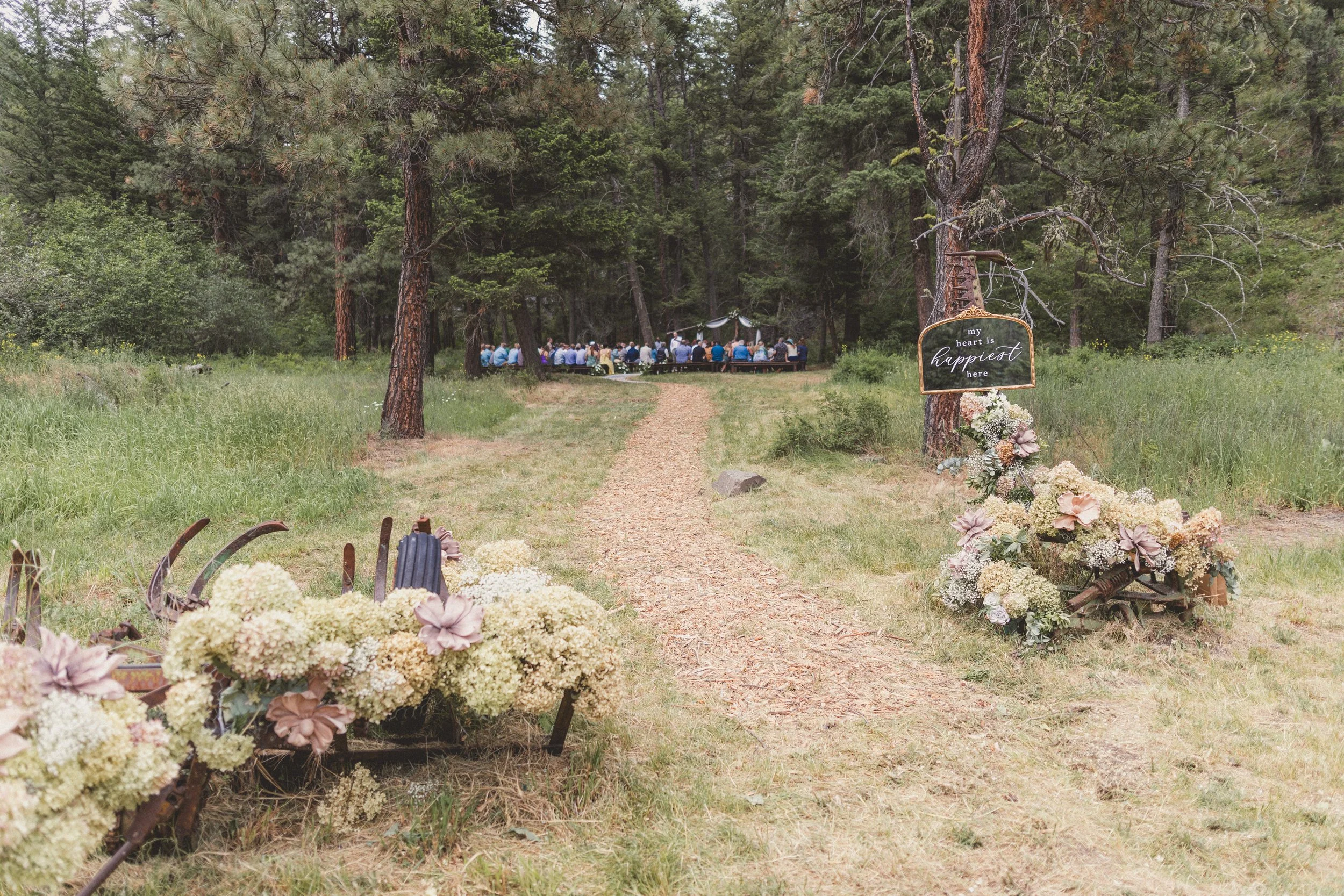 Outdoor wedding ceremony setup in a forest clearing, with rows of seats facing an altar, surrounded by trees and flowers, and a sign that reads 'my heart is happiest here'.