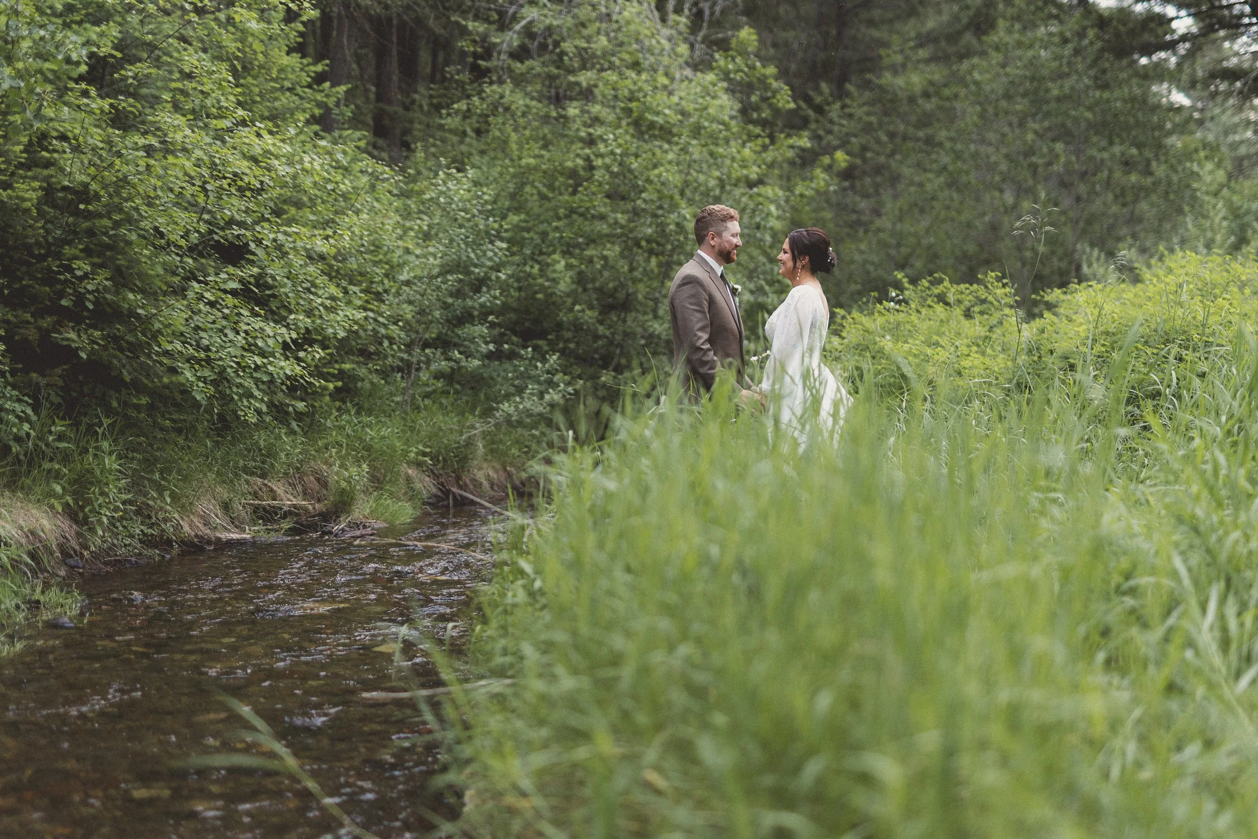 A bride and groom stand in a lush, green forest near a small stream, gazing into each other's eyes.