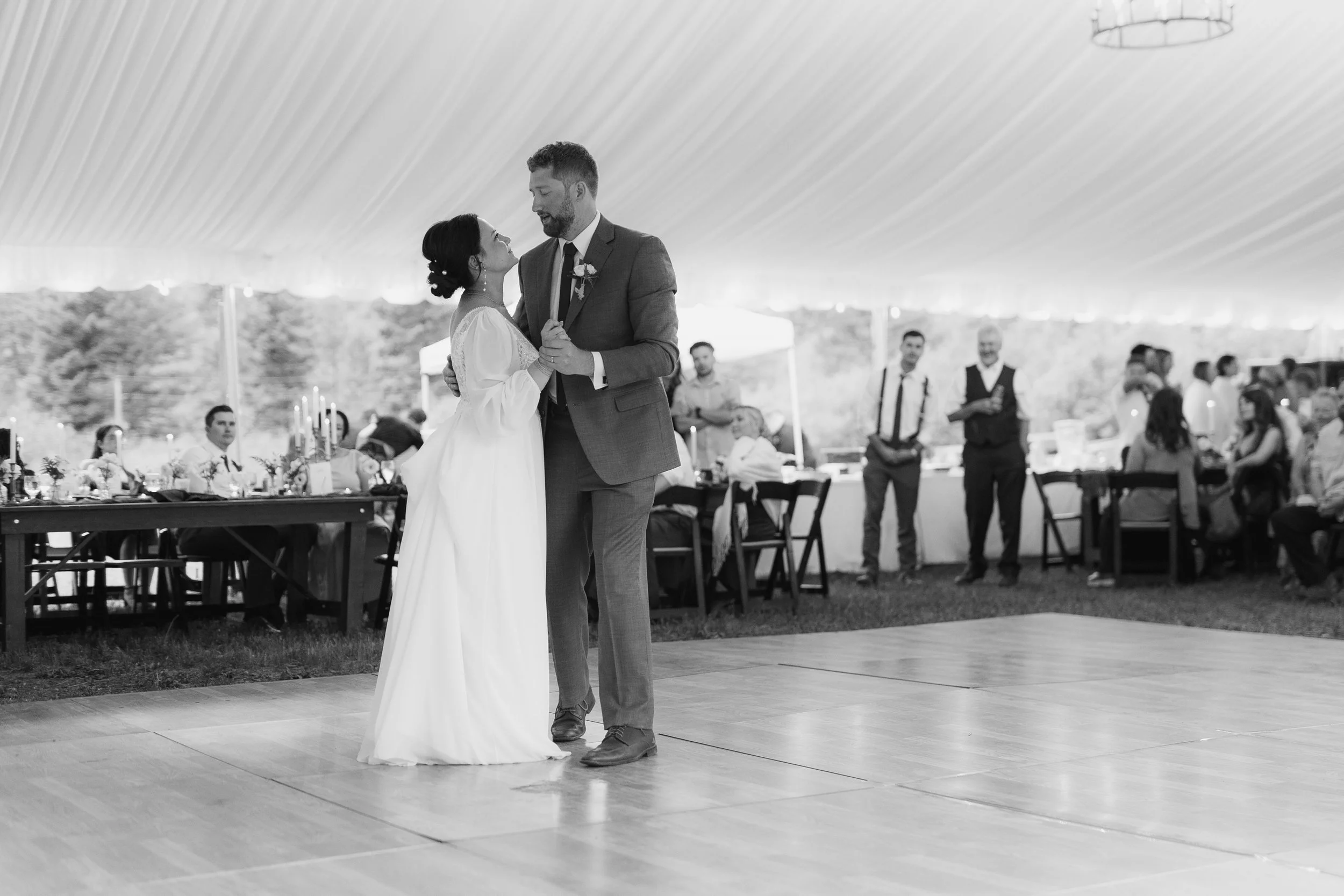 Black and white photo of a bride and groom dancing under a canopy at their wedding reception, with guests sitting and standing in the background.