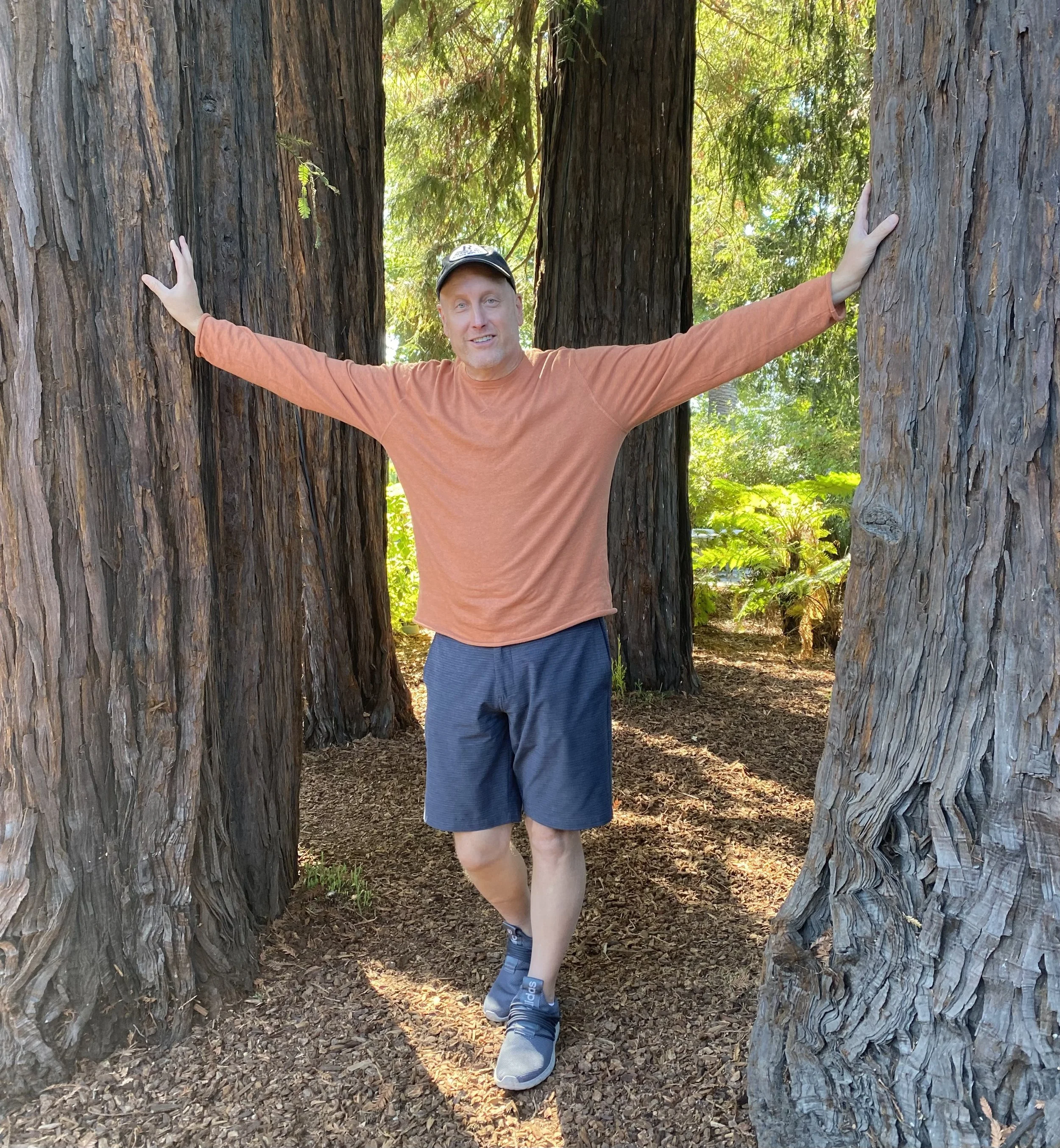A man in a long-sleeved orange shirt, blue shorts, and sneakers standing with arms outstretched between large redwood trees in a lush green forest.