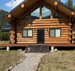 A log cabin with a front steps leading up to a black door, surrounded by grass and trees in the background.