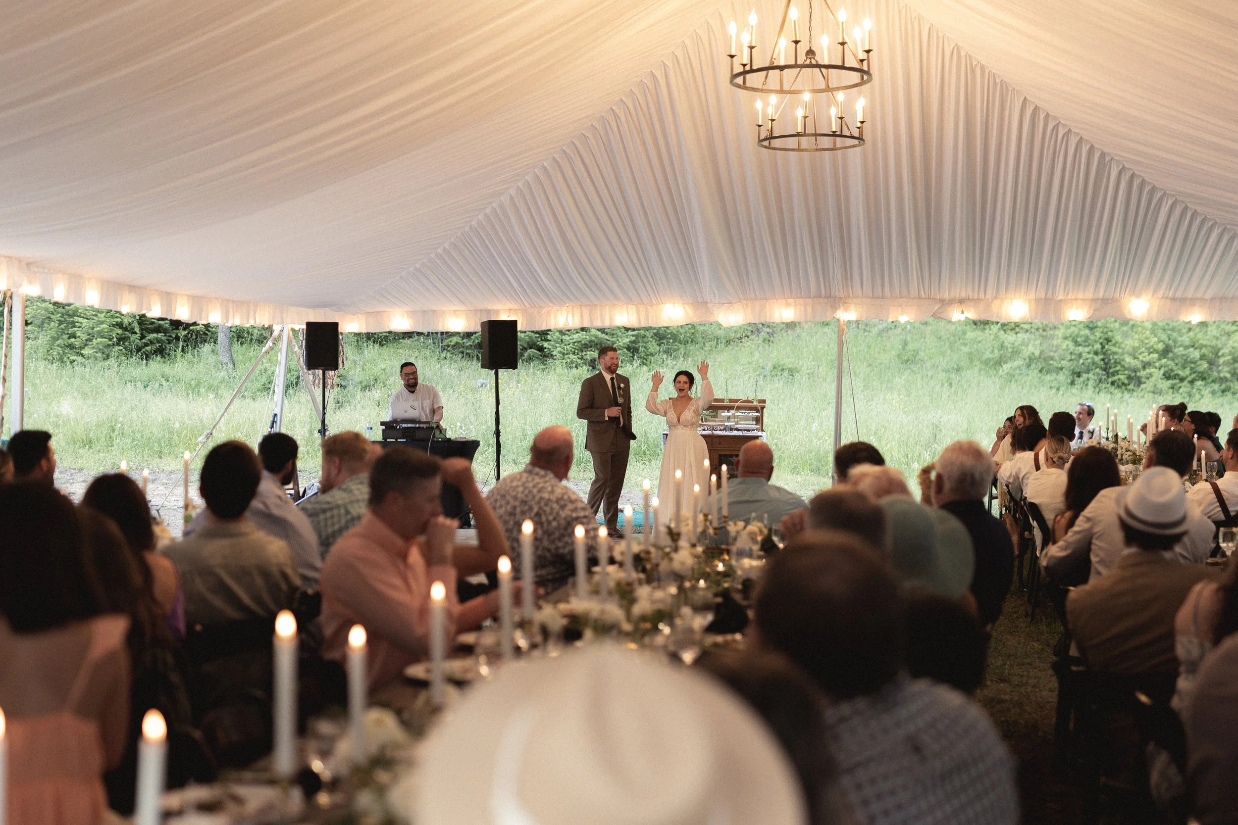 Wedding reception under a large outdoor tent with guests seated at decorated tables, a bride and groom standing at the front, artist playing music, and a lush green landscape outside.