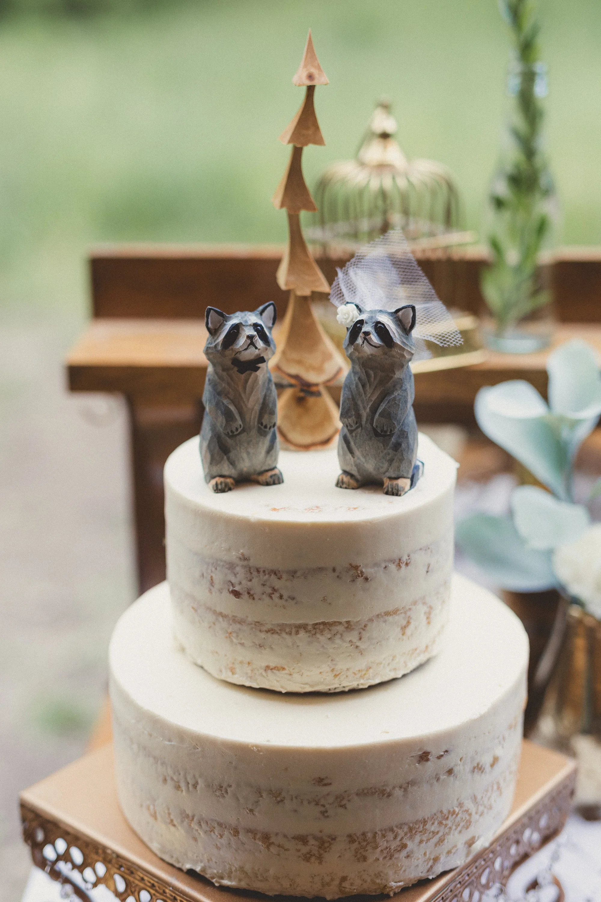 Two rustic tiered wedding cakes with raccoon figurines, one wearing a veil, on a decorative cake stand at a celebration.
