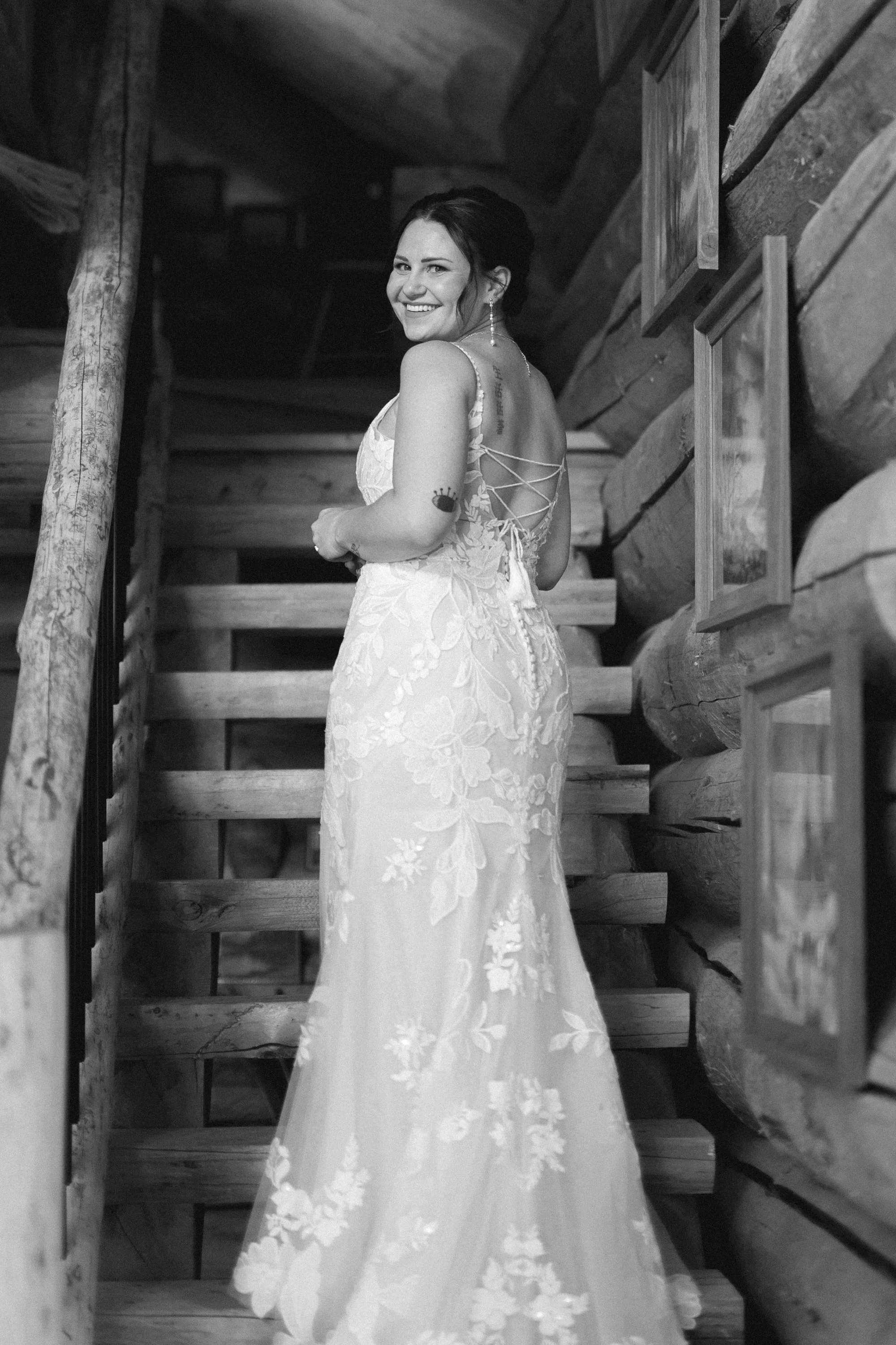 A smiling woman in a lace wedding dress with an open back standing on a wooden staircase in a log cabin.