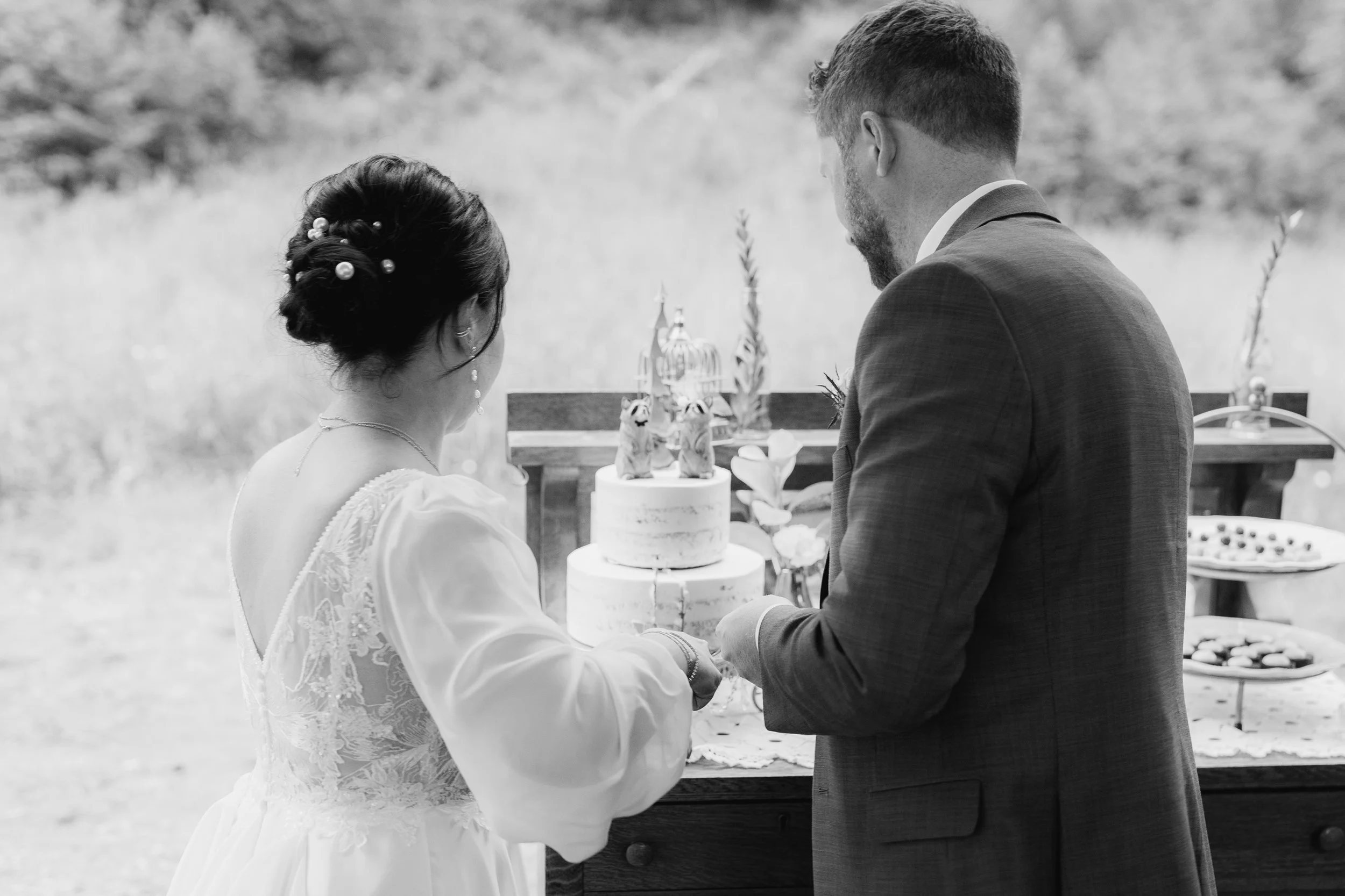 A bride and groom cutting a wedding cake outdoors, with desserts on a table behind them, in black and white.