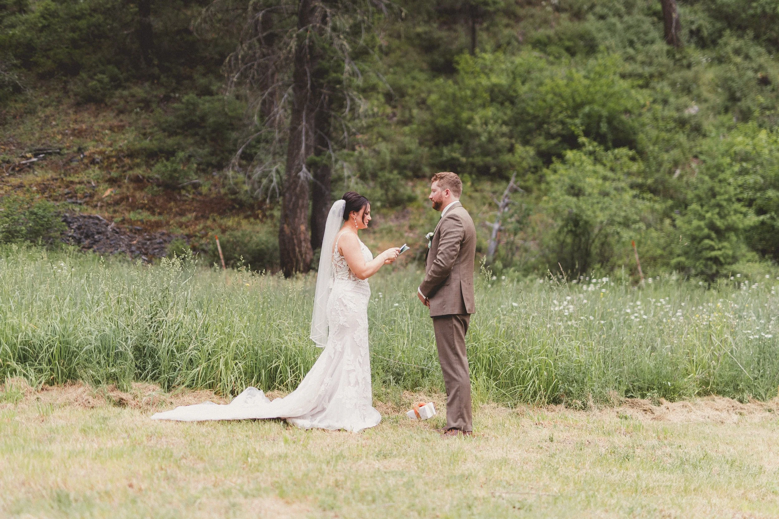 A bride and groom exchange vows outdoors in a green, natural setting with tall trees and grass, standing on a grassy area.