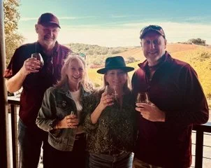 Four friends standing on a balcony with a scenic view, holding wine glasses and smiling.