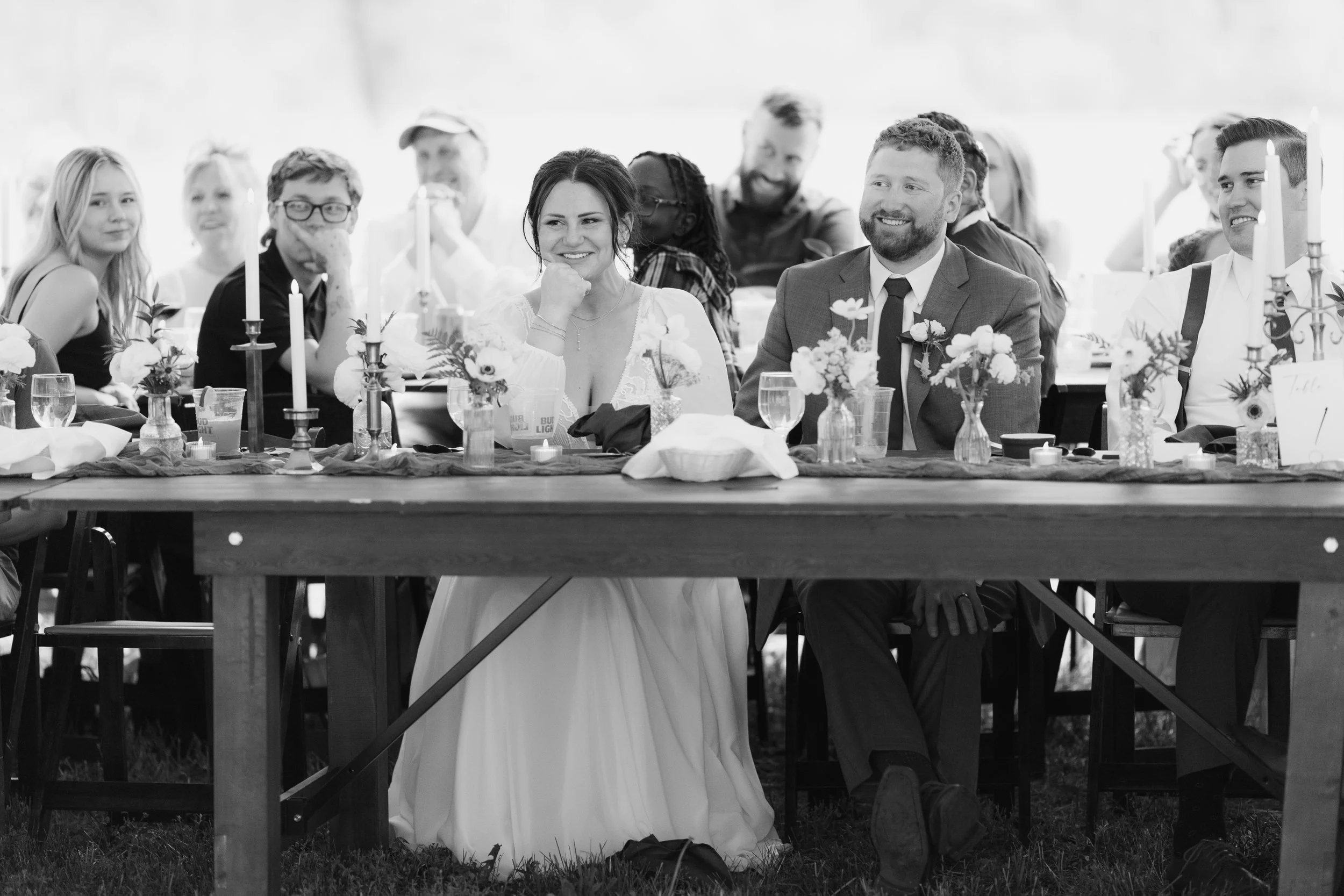 A group of wedding guests seated at a long table, with the bride and groom at the center. The bride is smiling and resting her chin on her hand, wearing a white dress. The groom, wearing a suit, is smiling. The table is decorated with flowers and can