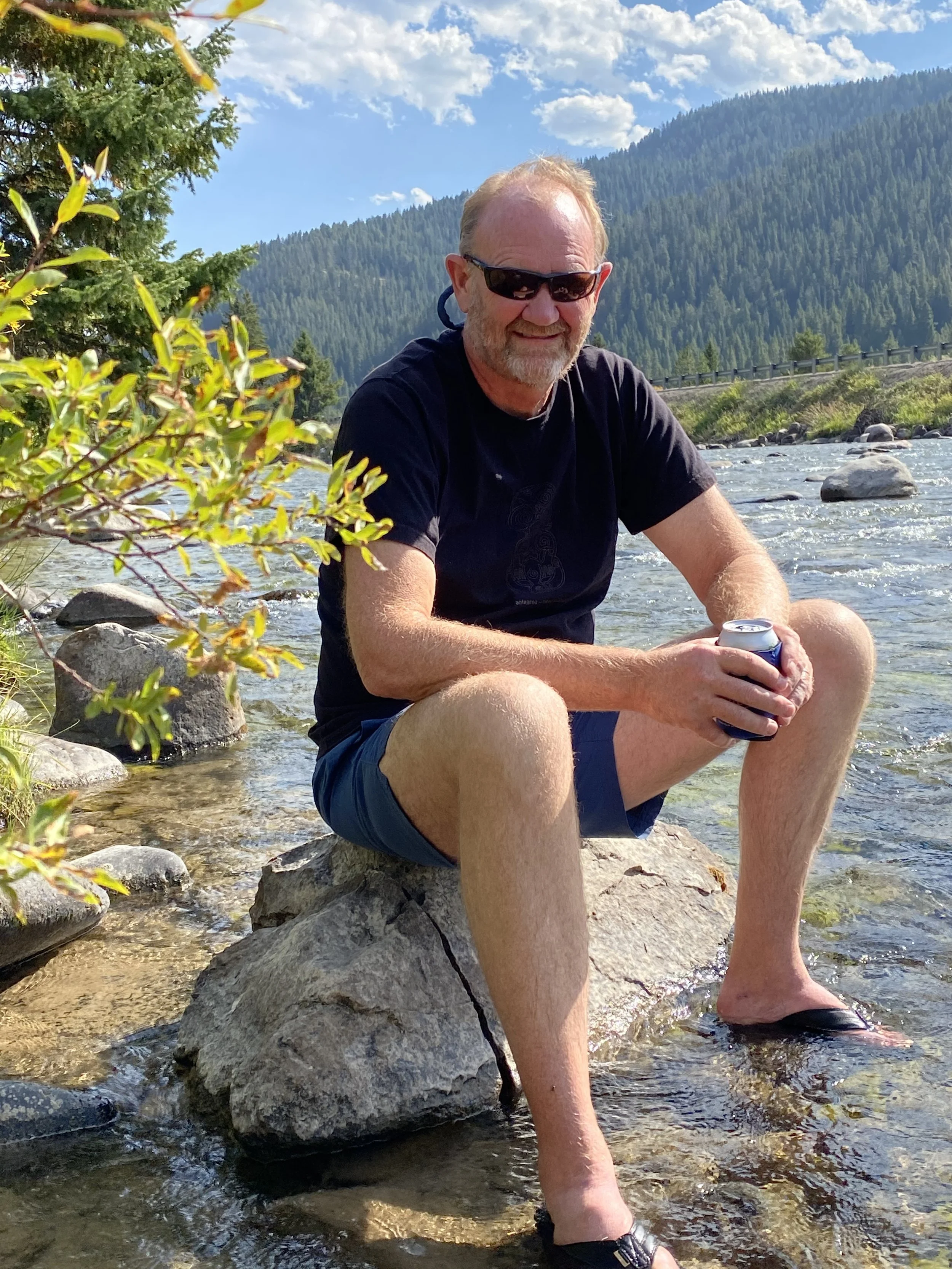 A man sitting on a rock in a river, holding a can. He is wearing sunglasses, a black t-shirt, shorts, and sandals. There are trees, mountains, and a partly cloudy sky in the background.