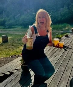 A woman sitting outdoors on a wooden platform, smiling and holding a bottle and a glass of beer, with a scenic green landscape and mountains in the background.