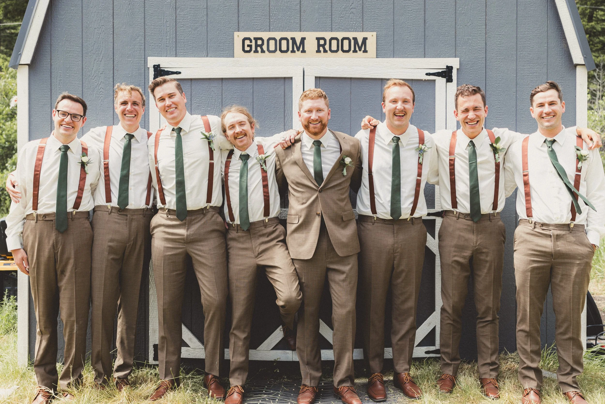 A group of nine men standing arm-in-arm in front of a barn with a sign that reads "GROOM ROOM," dressed in wedding attire with suspenders and ties, smiling for the camera.