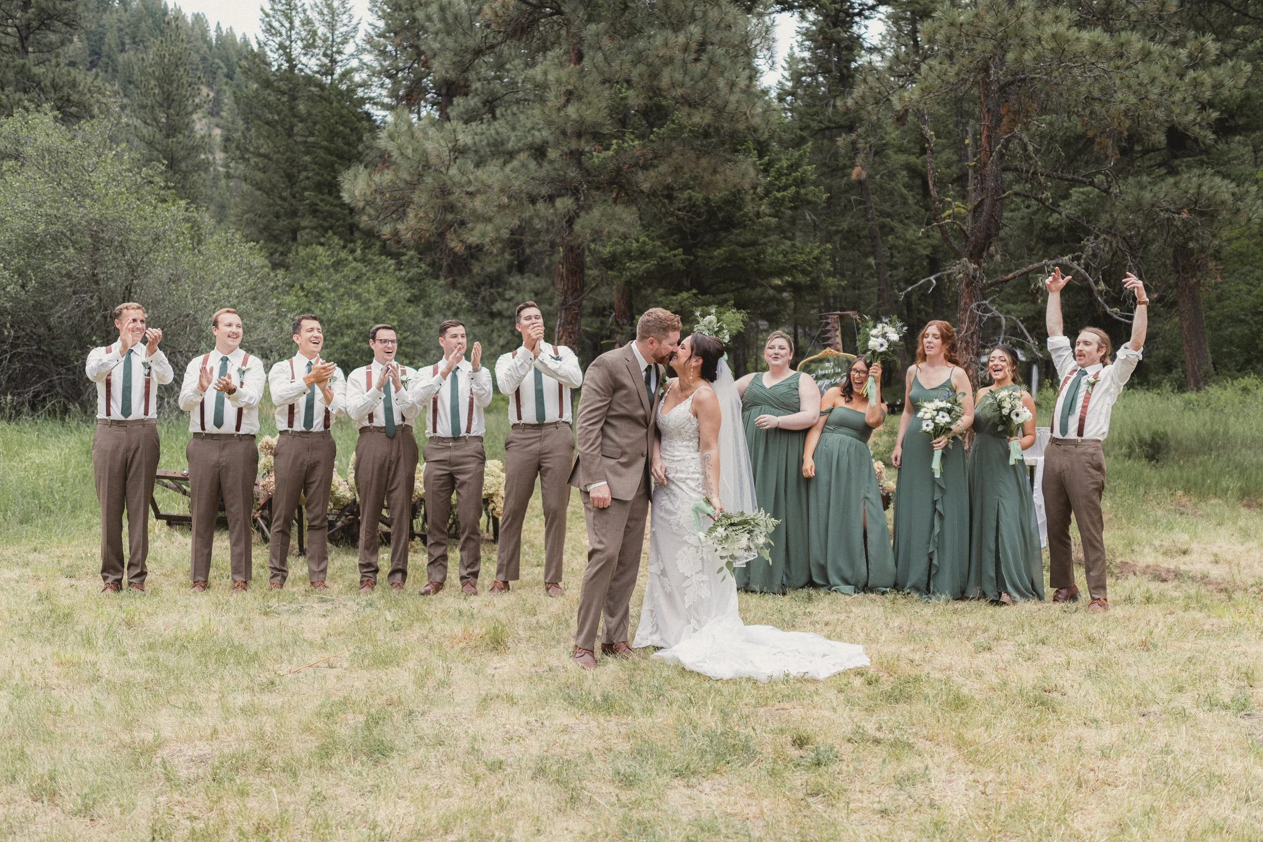 A wedding ceremony outdoors with the bride and groom kissing. The wedding party includes six groomsmen and six bridesmaids, all standing in front of a forested area. The bridesmaids are in green dresses, and the groomsmen are in beige pants, white sh