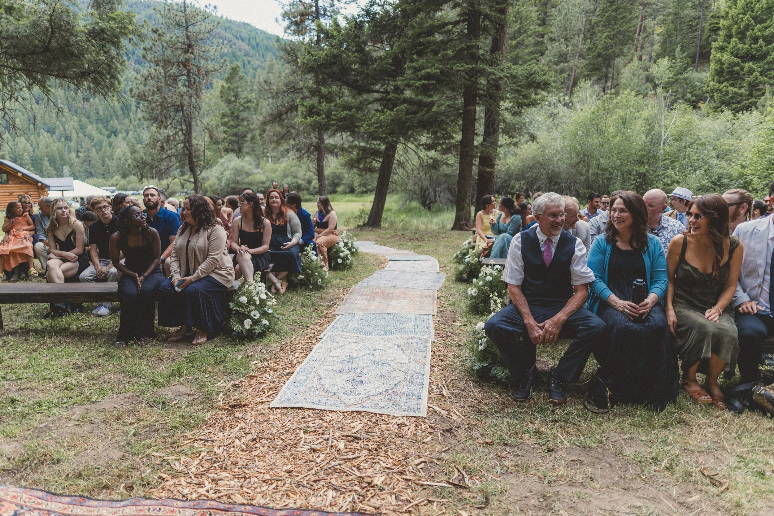 Guests seated outdoors on benches and chairs, attending a wedding ceremony in a forested area with mountains in the background, decorated with white flowers along a walkway.