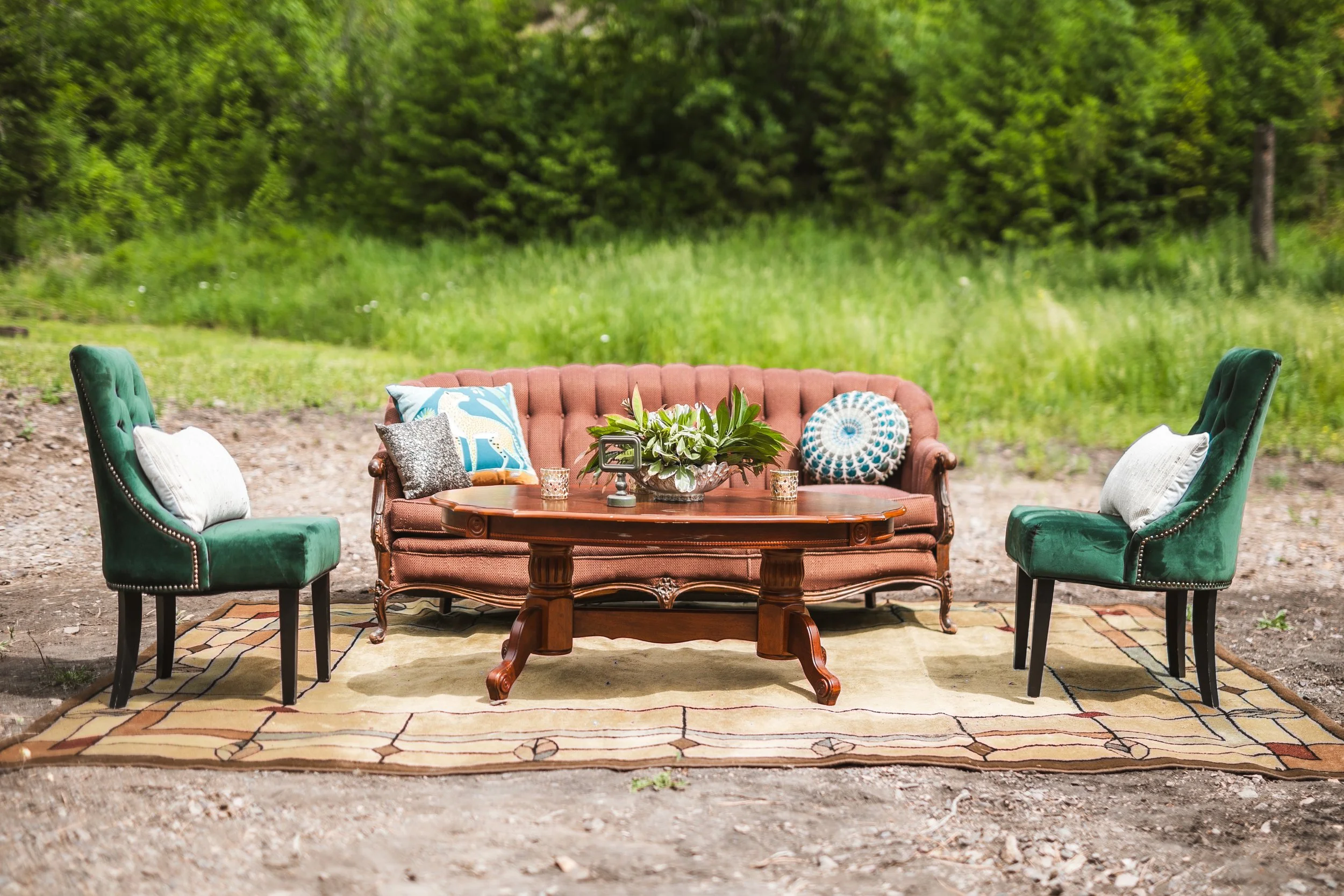 Outdoor furniture arranged on a rug in a natural setting with green grass and trees in the background. The setup includes a vintage-style sofa with decorative pillows, a wooden coffee table with a plant centerpiece and candles, and two green velvet a