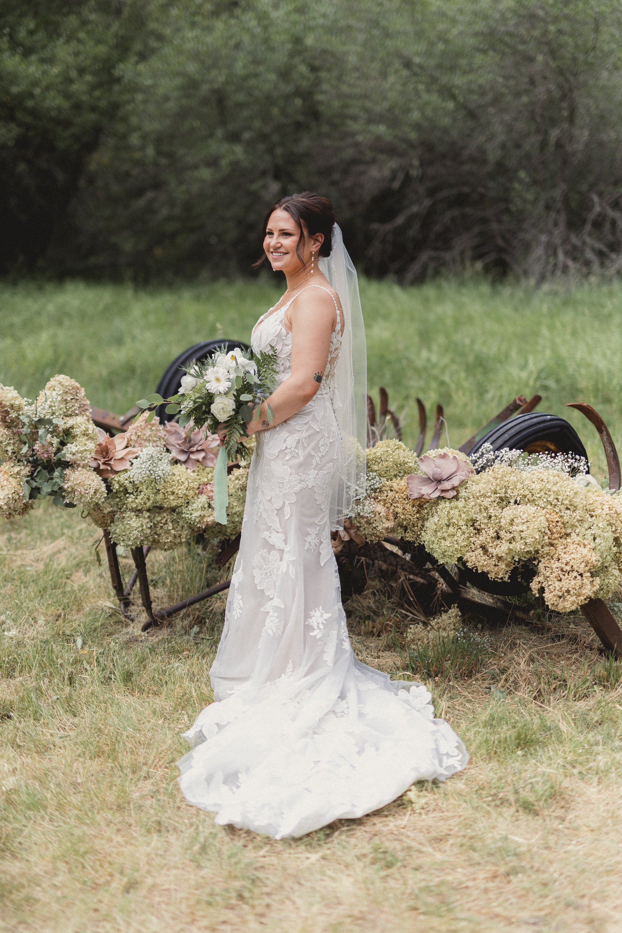 A bride in a white lace wedding dress with a veil, holding a bouquet, standing outdoors on grass with a vintage wagon decorated with large flowers and greenery in the background.