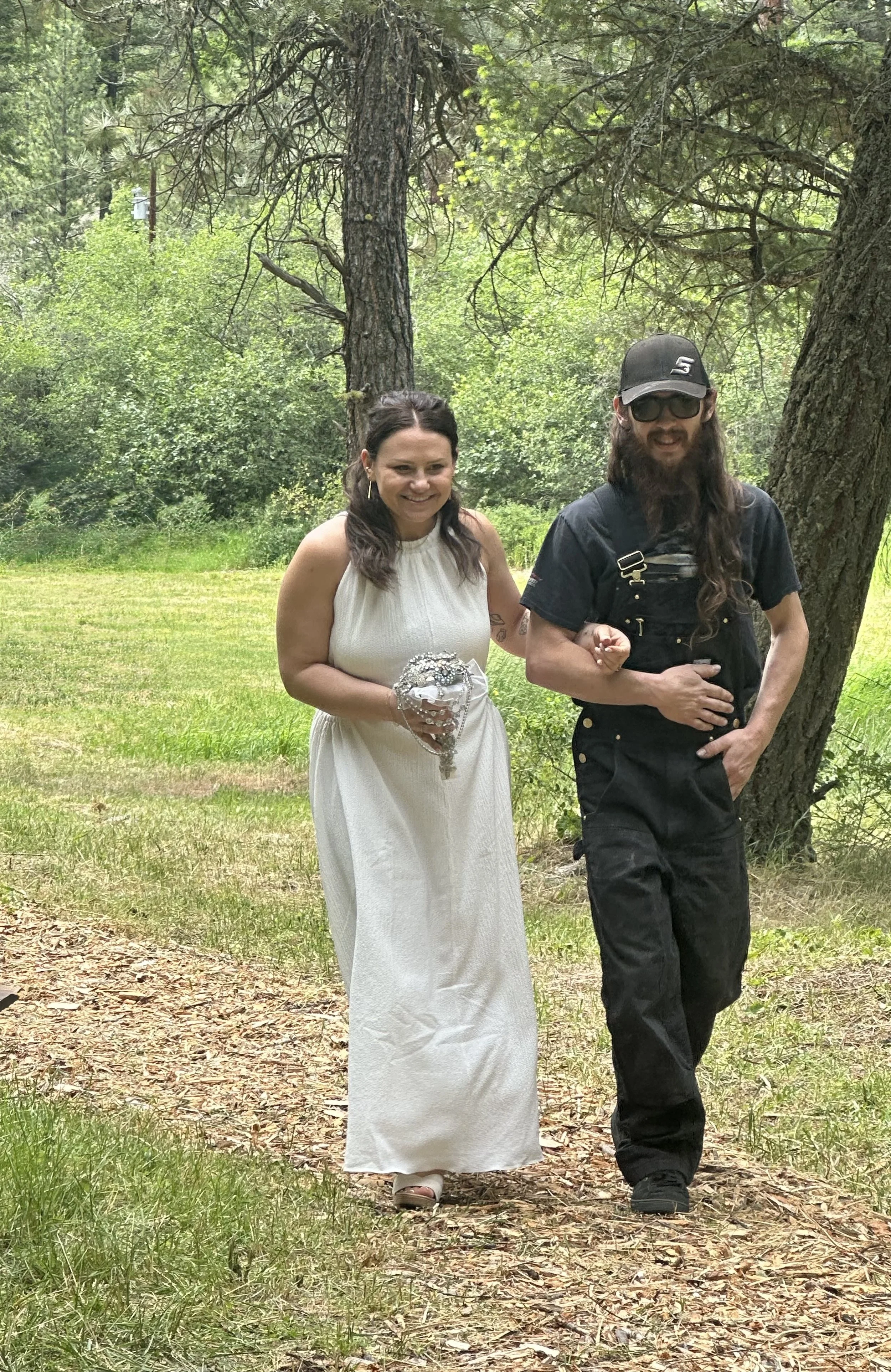 A woman in a white dress holding a small bouquet walking arm-in-arm with a man in black overalls walking outdoors on a wooded trail with trees and greenery in the background.