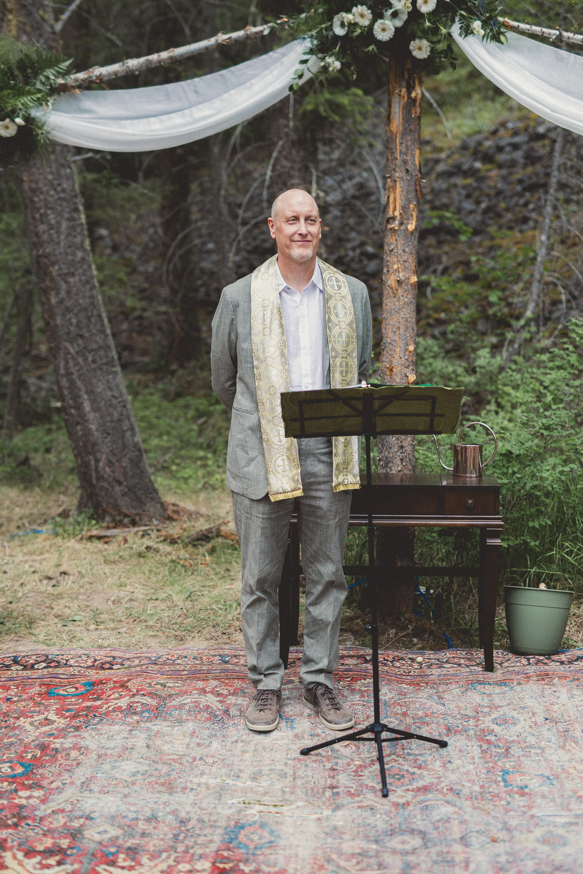 A man in a gray suit with a white shirt and patterned stole stands outdoors under a decorated wooden arch, next to a music stand and a small table with a copper watering can, conducting a ceremony or service in a forest setting.