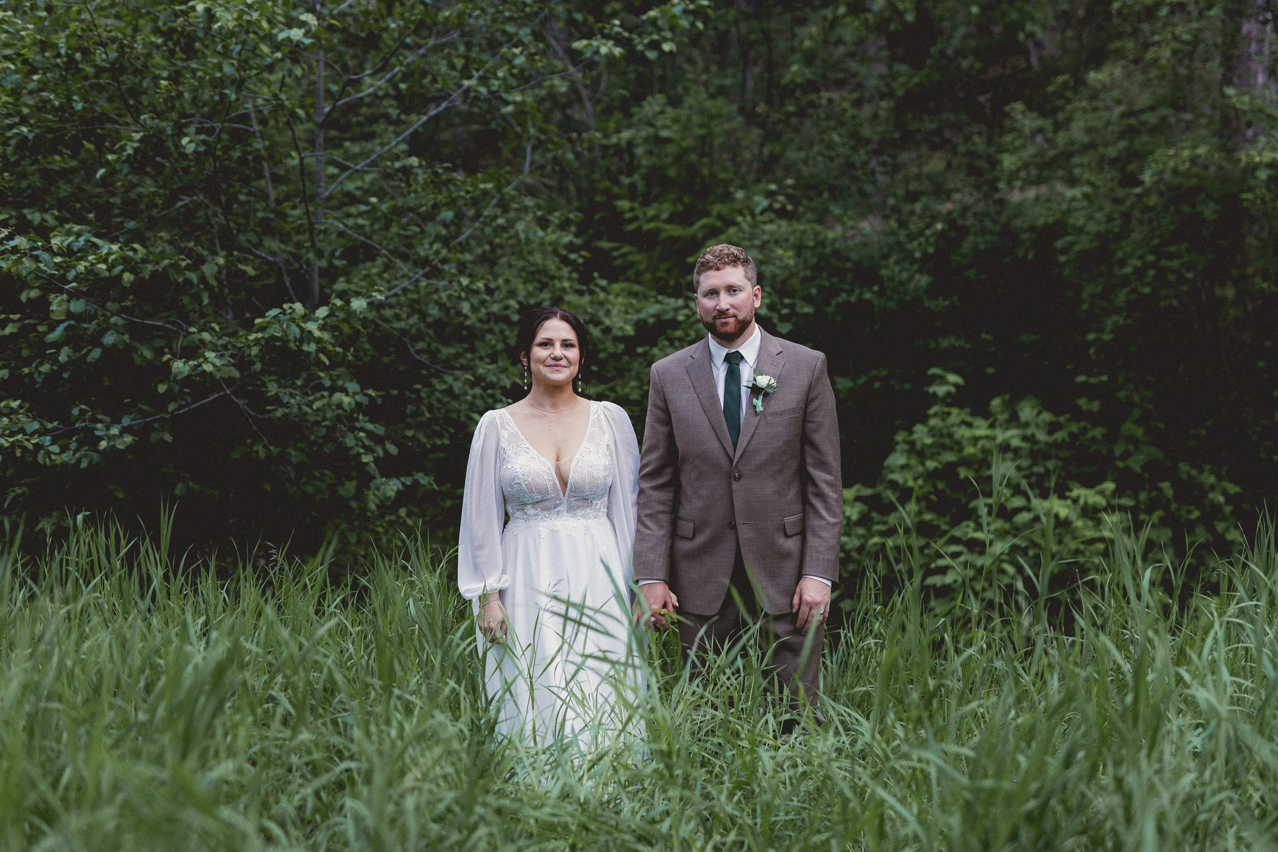 A bride and groom standing in tall grass outdoors with dense greenery and trees in the background, during their wedding photos.