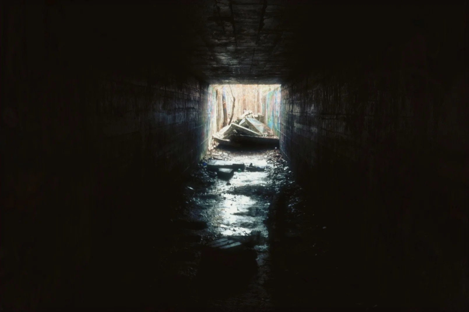 Dark tunnel with debris and water, leading to a bright opening outside where broken objects are visible.