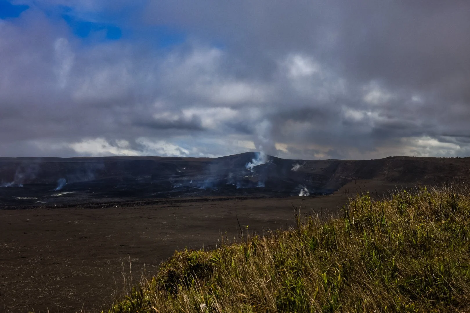 A volcanic landscape with dark, smoky clouds overhead, a volcano emitting smoke and ash in the distance, and patches of grass in the foreground.