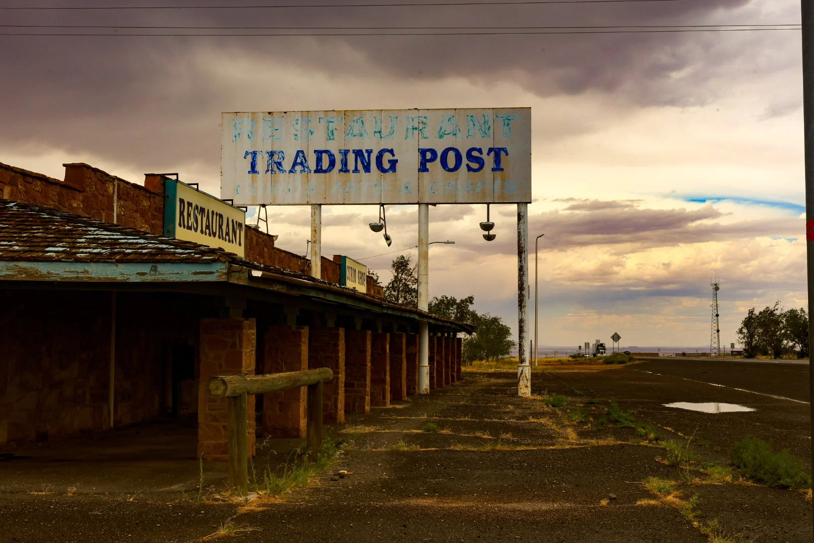 An old, weathered sign on a roadside advertising a restaurant and trading post, with cloudy skies and a distant horizon in the background.