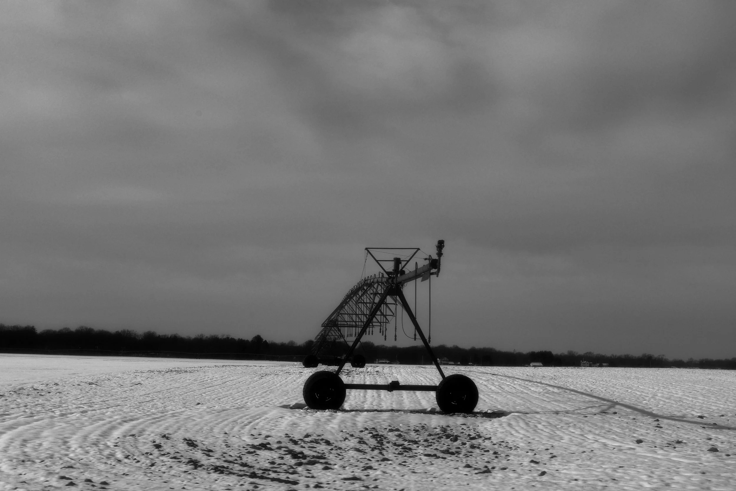 A black and white photo of a large, industrial water sprinkler system in a snow-covered field, with a cloudy sky overhead.