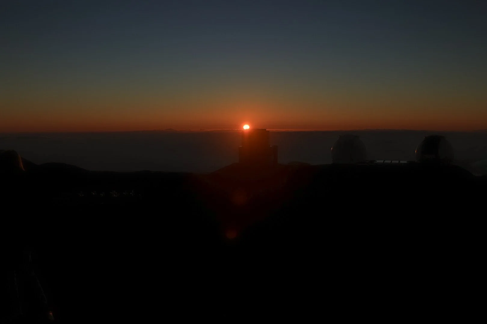 Sunset seen from an observatory with multiple telescope domes, with the sun near the horizon and a colorful sky.