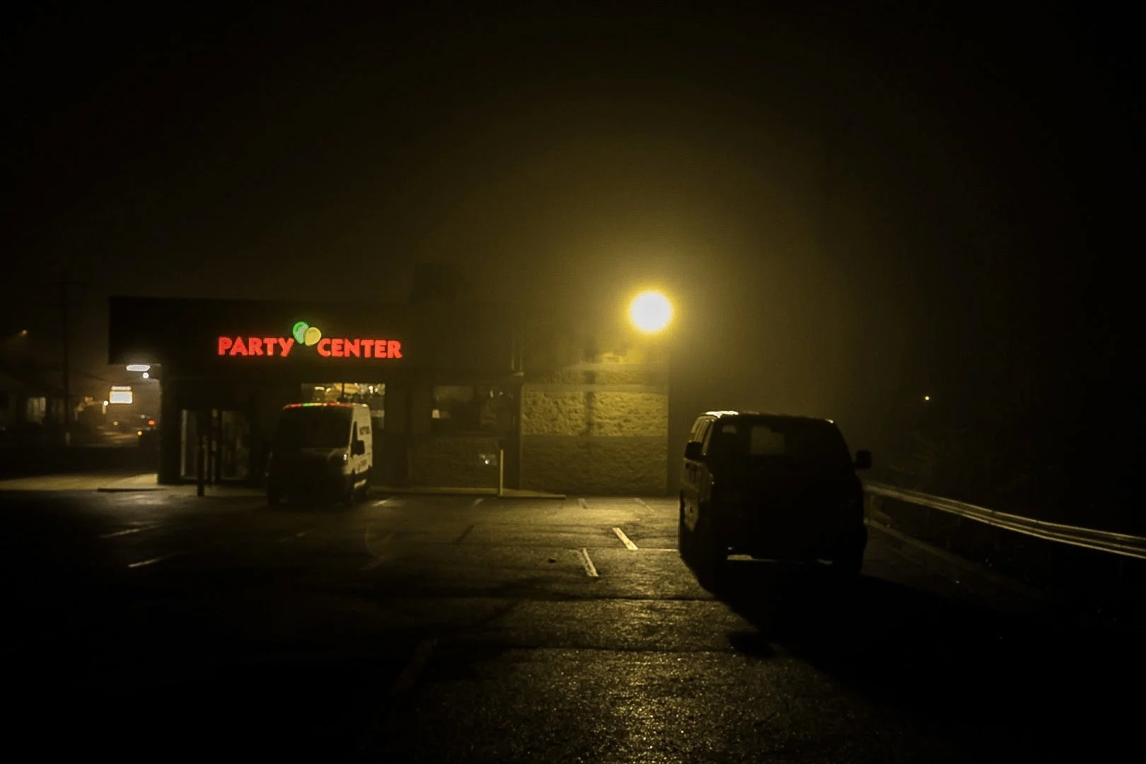 Nighttime scene of a parking lot in foggy weather with a neon sign reading 'PARTY CENTER' and a bright streetlight illuminating the area.