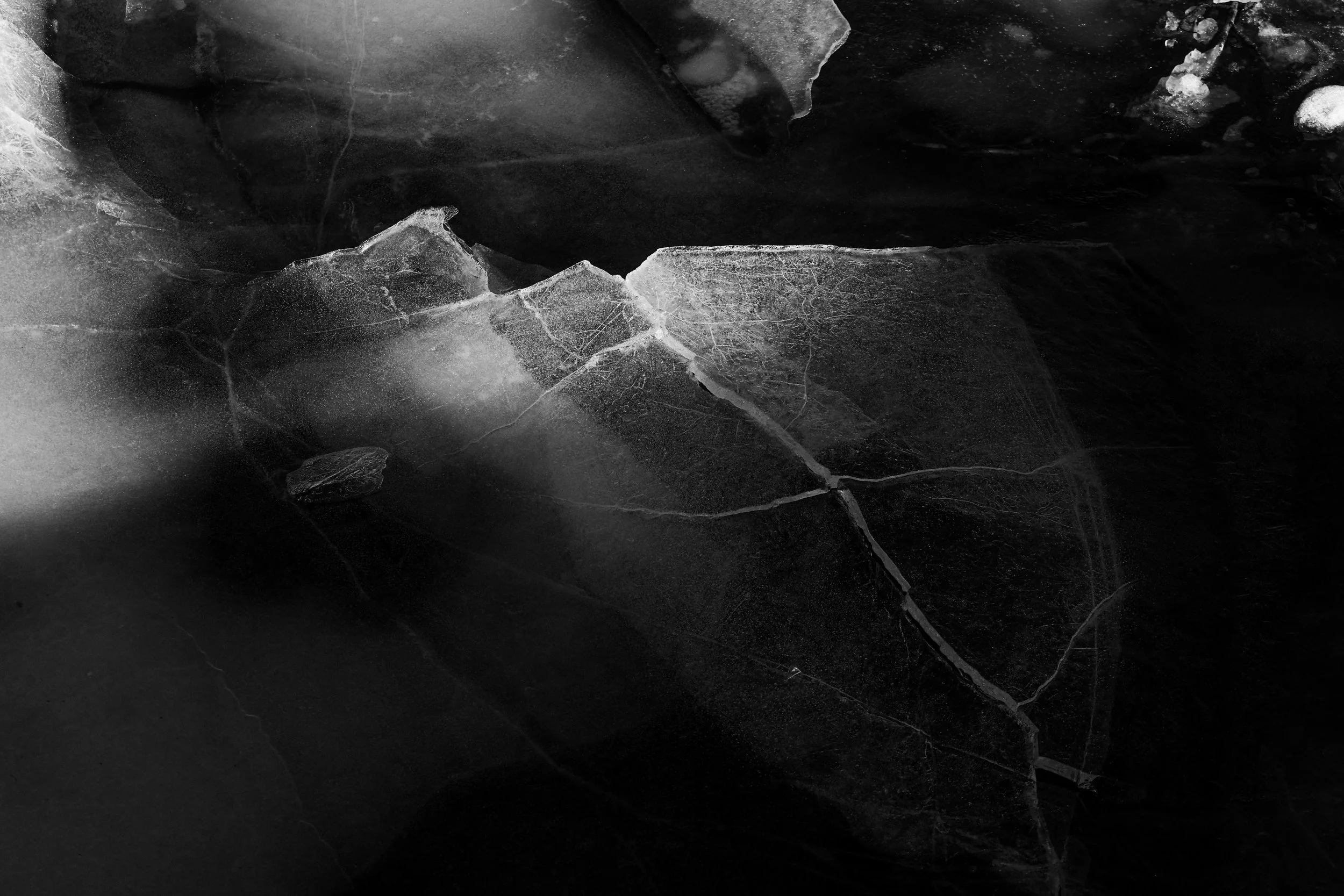 Close-up of cracked and broken ice on a dark surface, showing jagged edges and cracks in black and white.