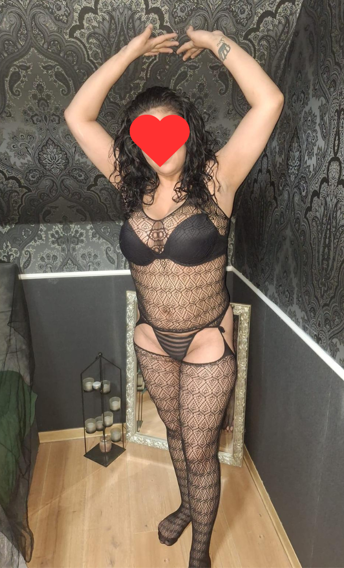 Frau in schwarzem, durchsichtigen Spitzen-Body mit Strumpfhose, posiert vor einem Spiegel in einem Raum mit schwarz gemusterten Tapeten. Gesicht durch einen roten Herz-Emoji verdeckt.