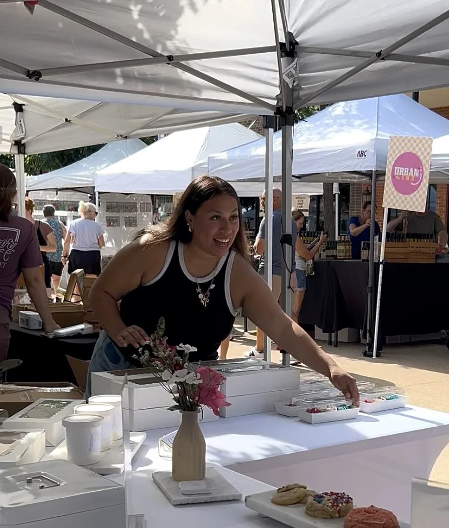 A woman at an outdoor market stall displays baked goods and small containers under a white canopy.