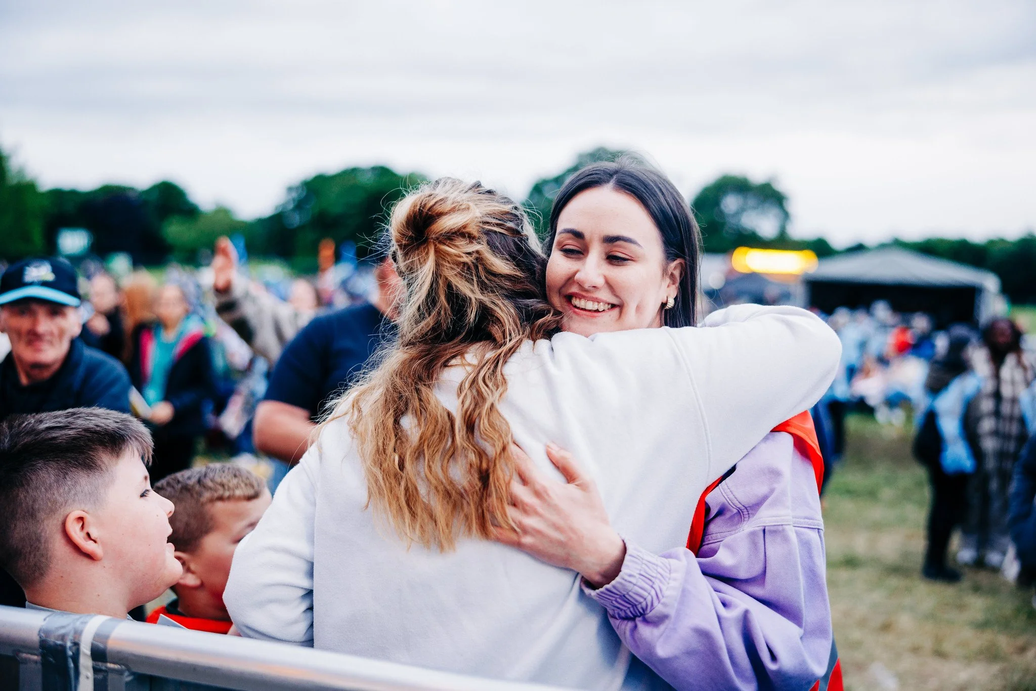 Two women hugging at an outdoor event, smiling, with a crowd of people in the background.