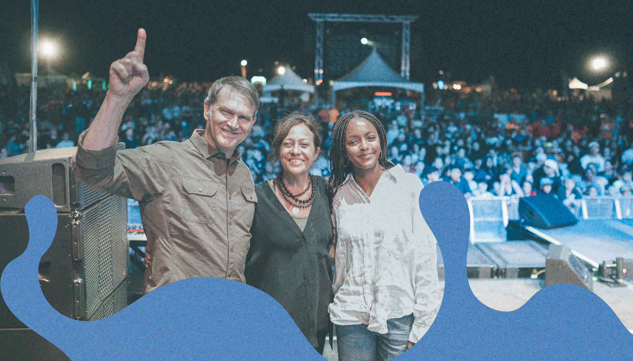 Three people stand together on a stage at an outdoor concert or event at night, with a large crowd in the background.