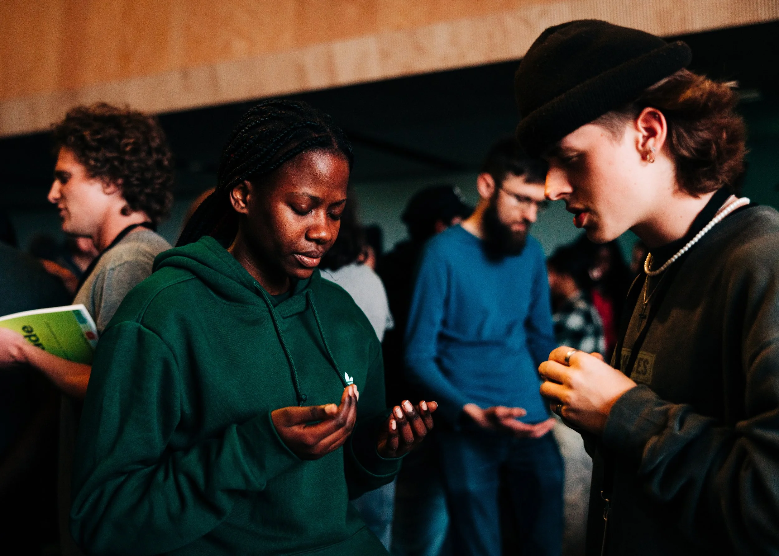 A group of young people standing together, with two of them in the foreground looking down and holding their hands, possibly engaged in prayer or a moment of reflection, in an indoor setting.