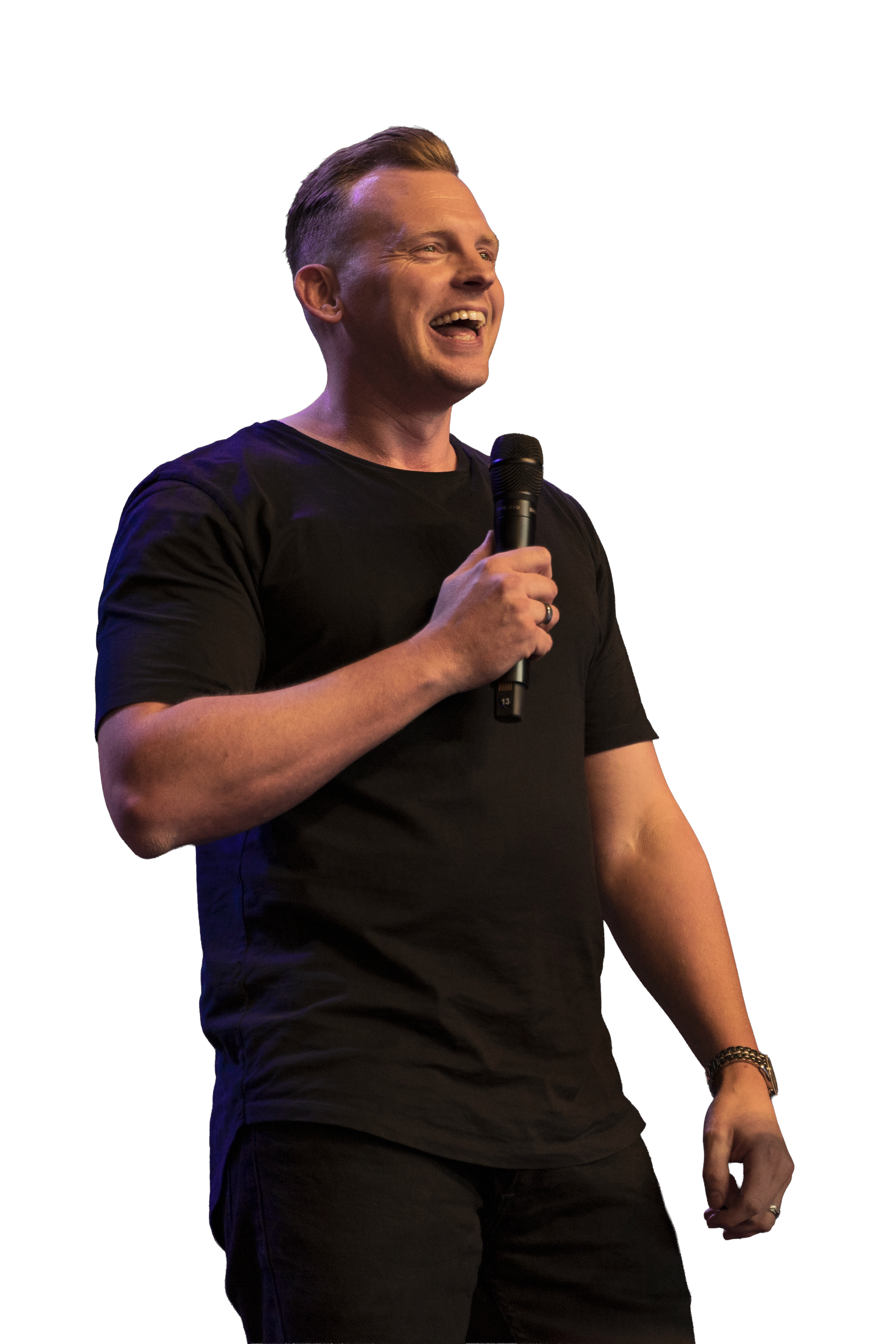 A man with short hair wearing a black t-shirt holding a microphone, smiling on stage with colorful lighting in the background.