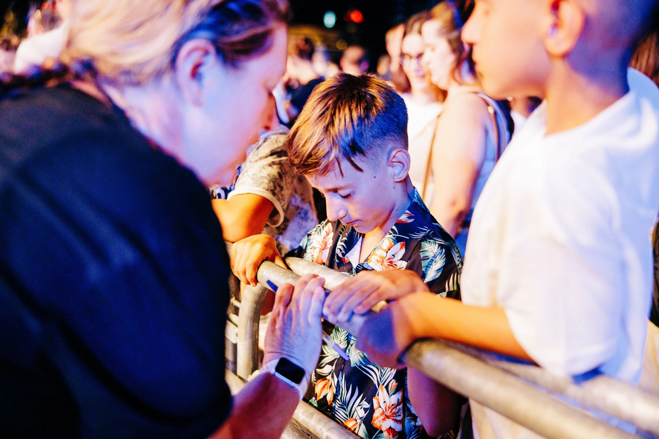 People are standing around a railing, with one young boy in the center holding the railing and bowing his head. An older woman is in front of him, possibly praying or blessing. Several others are visible in the background, some smiling, all engaged in a communal moment.