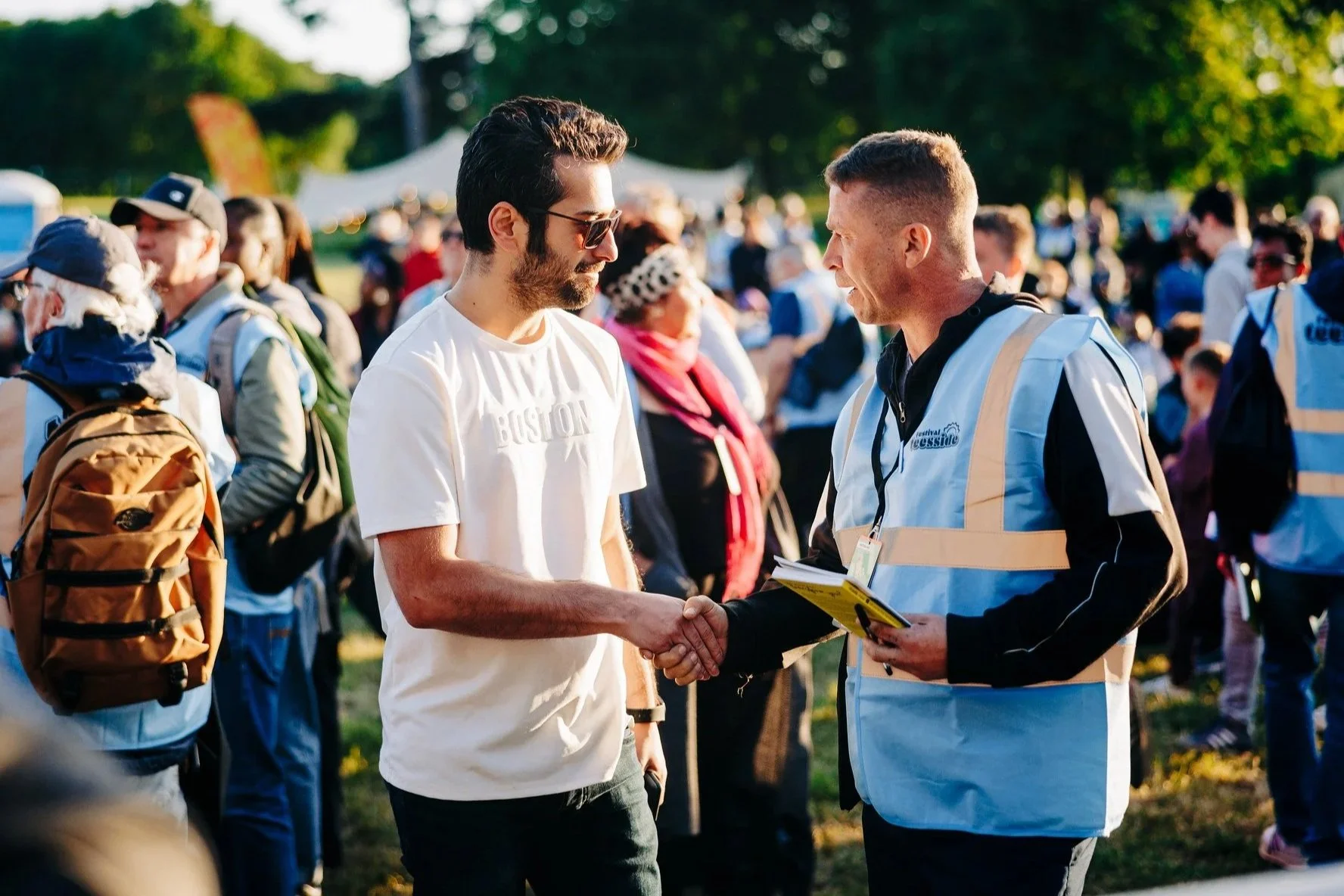 People are standing around a railing, with one young boy in the center holding the railing and bowing his head. An older woman is in front of him, possibly praying or blessing. Several others are visible in the background, some smiling, all engaged in a communal moment.