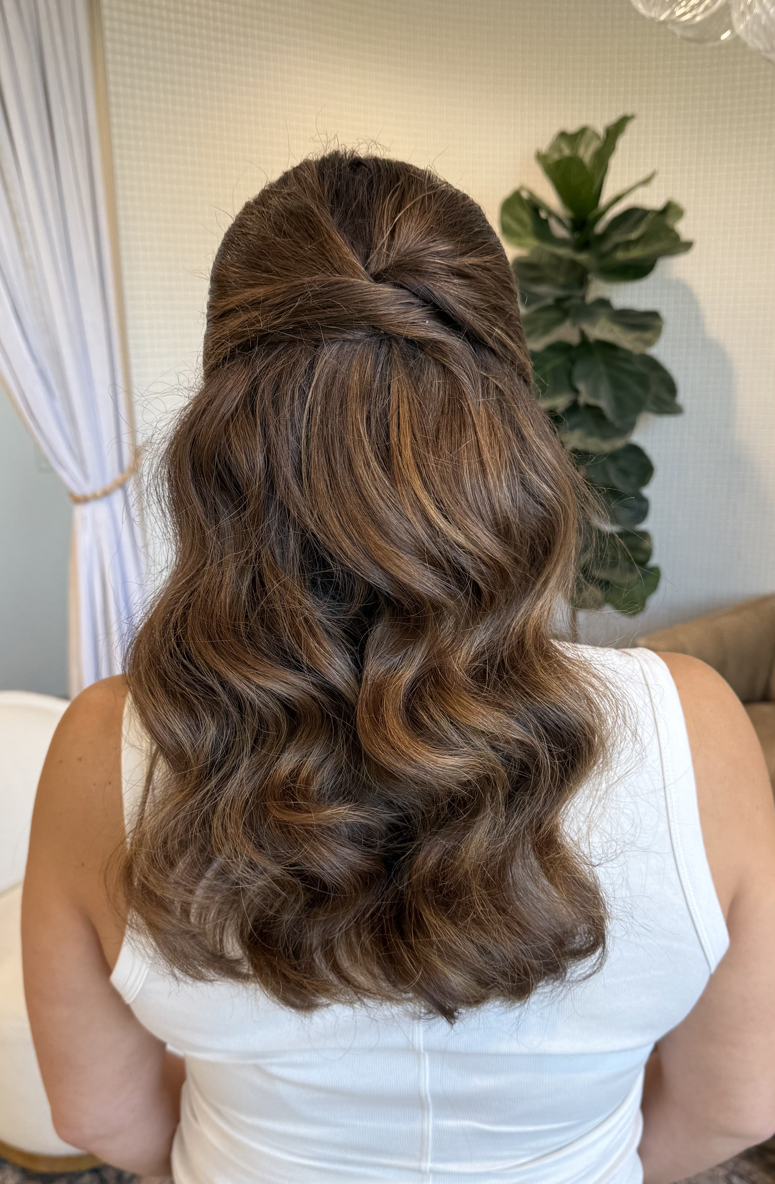 Back view of a woman with styled, wavy brown hair with highlights, wearing a white sleeveless top, standing indoors near a plant and curtain.