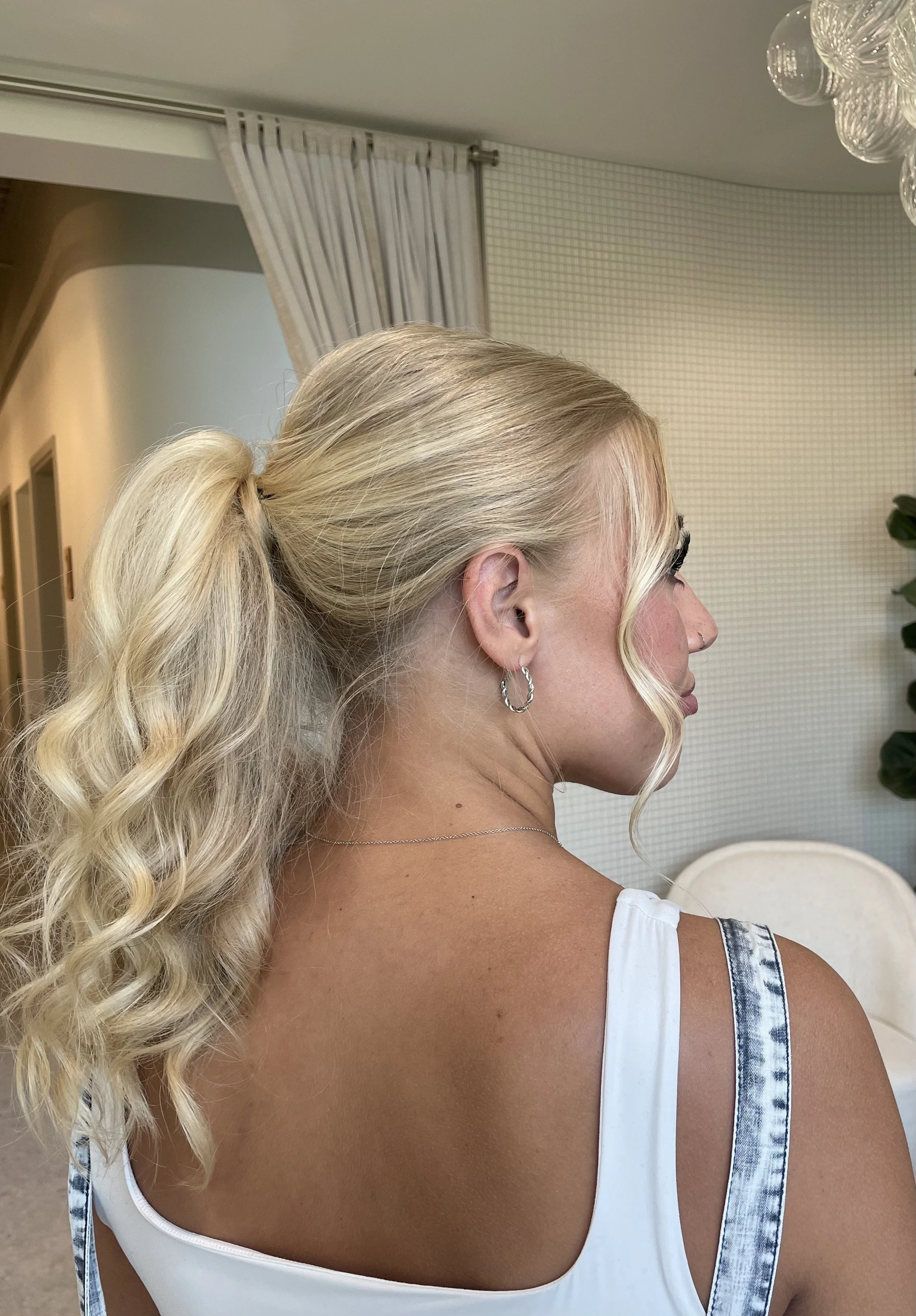 A woman with long, wavy blonde hair tied in a high ponytail, wearing earrings and a tank top, shown from the back and in profile.