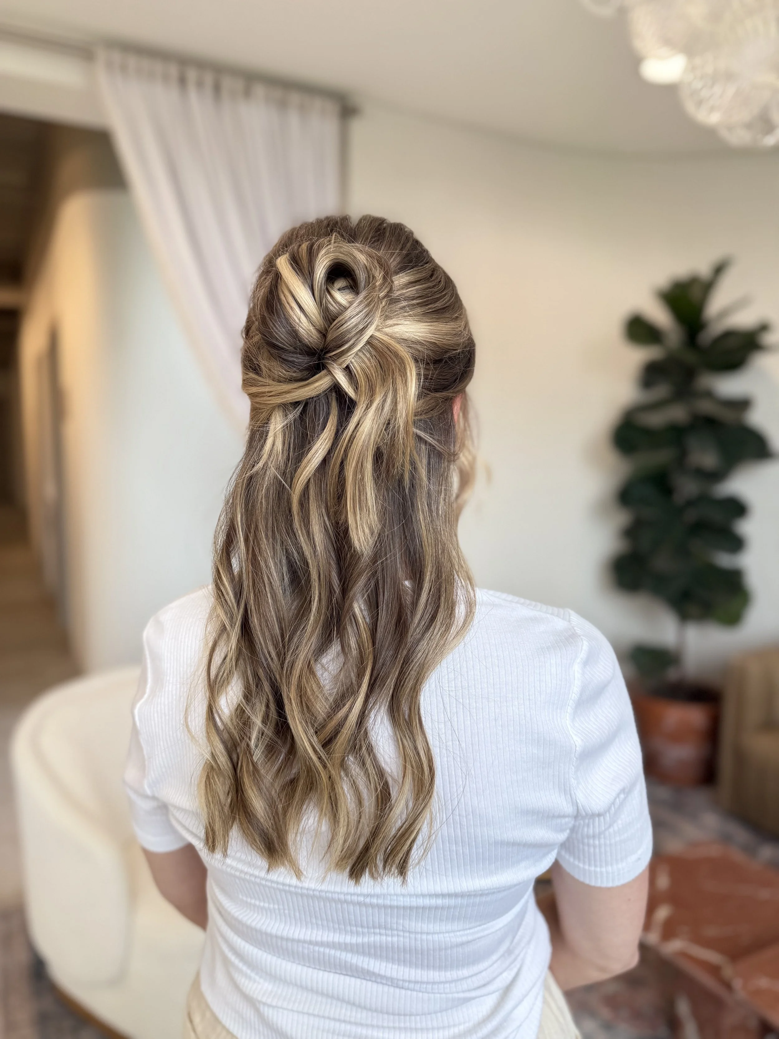 Back view of a woman with wavy blonde hair styled in an elegant updo with loose waves, sitting in a room with neutral decor.
