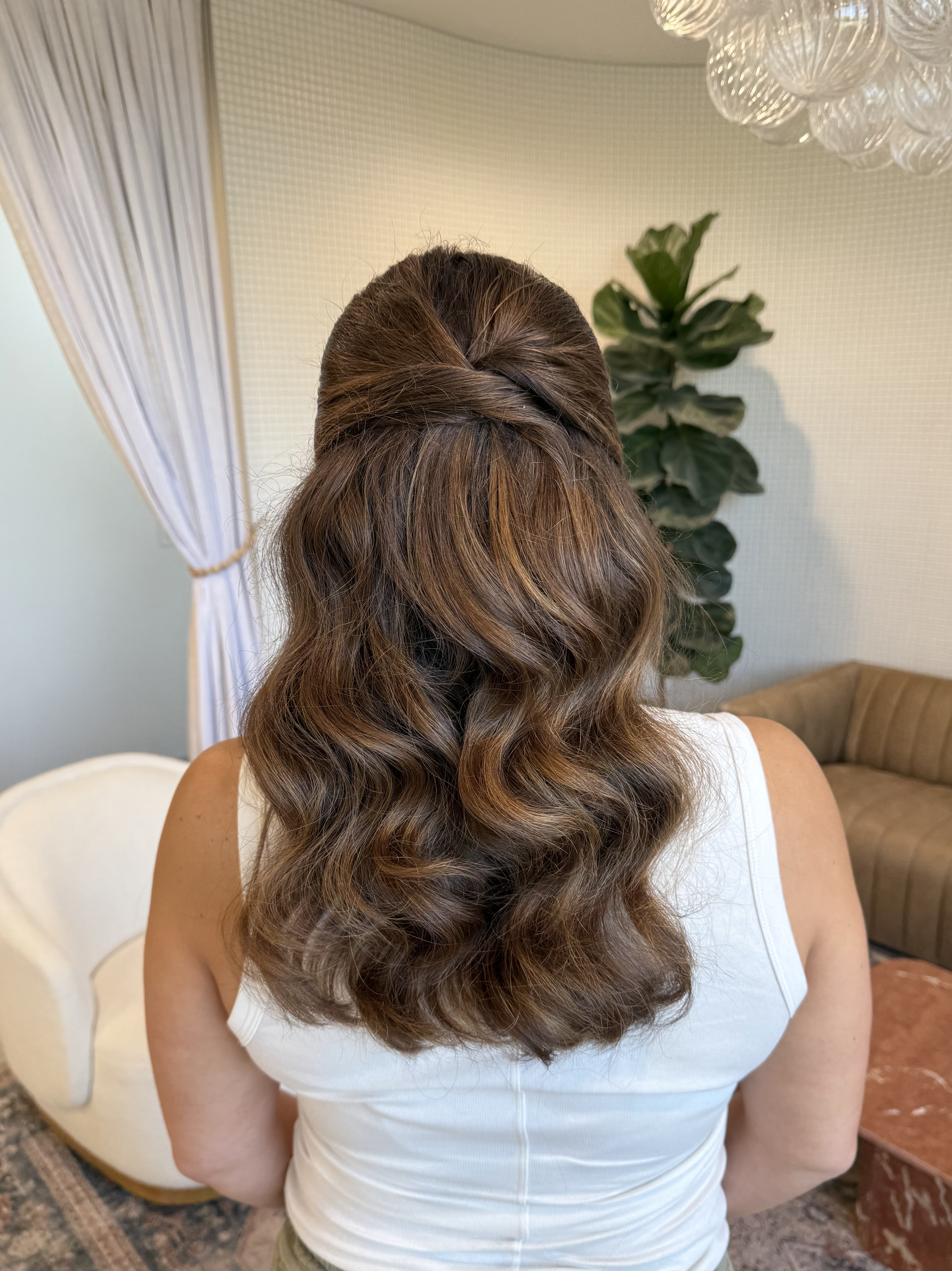 Woman with wavy brown hair styled in a half-updo, seen from the back, wearing a white sleeveless top indoors with a green plant and beige furniture in the background.