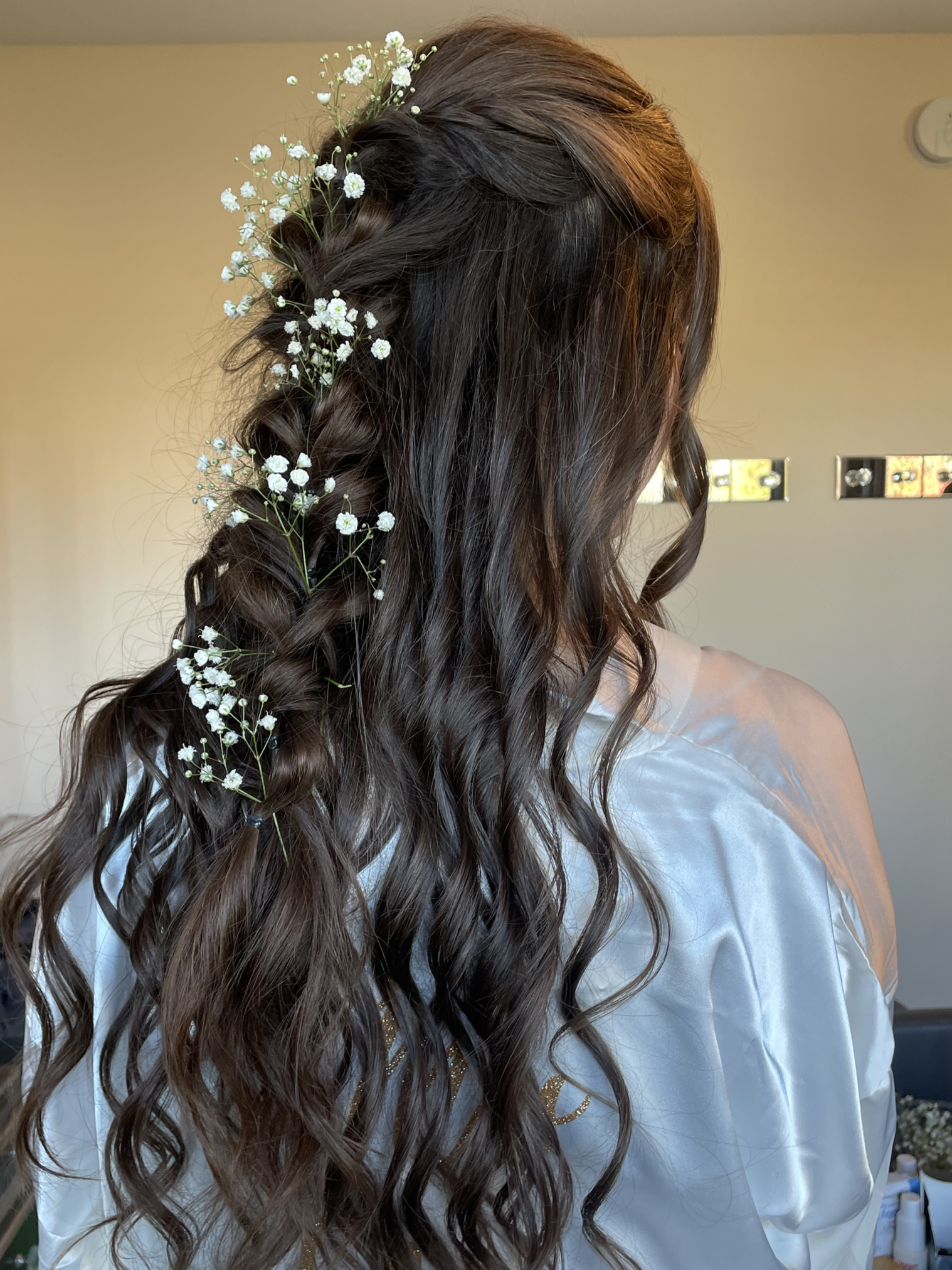 Side view of a woman with long, wavy brown hair decorated with small white flowers, wearing a white satin robe.