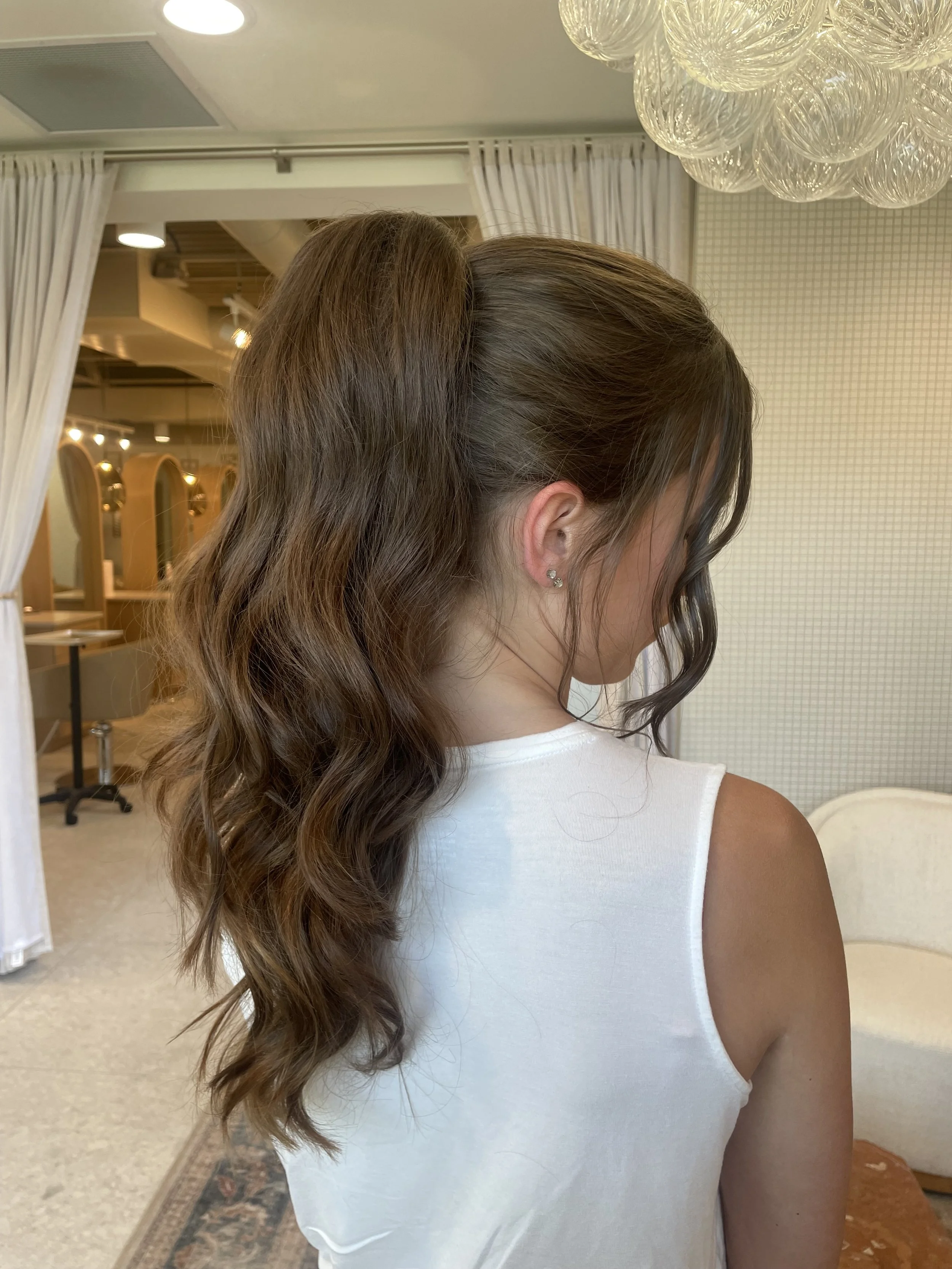 Back view of a woman with long wavy brown hair in a salon or salon-like setting, wearing a white sleeveless top.