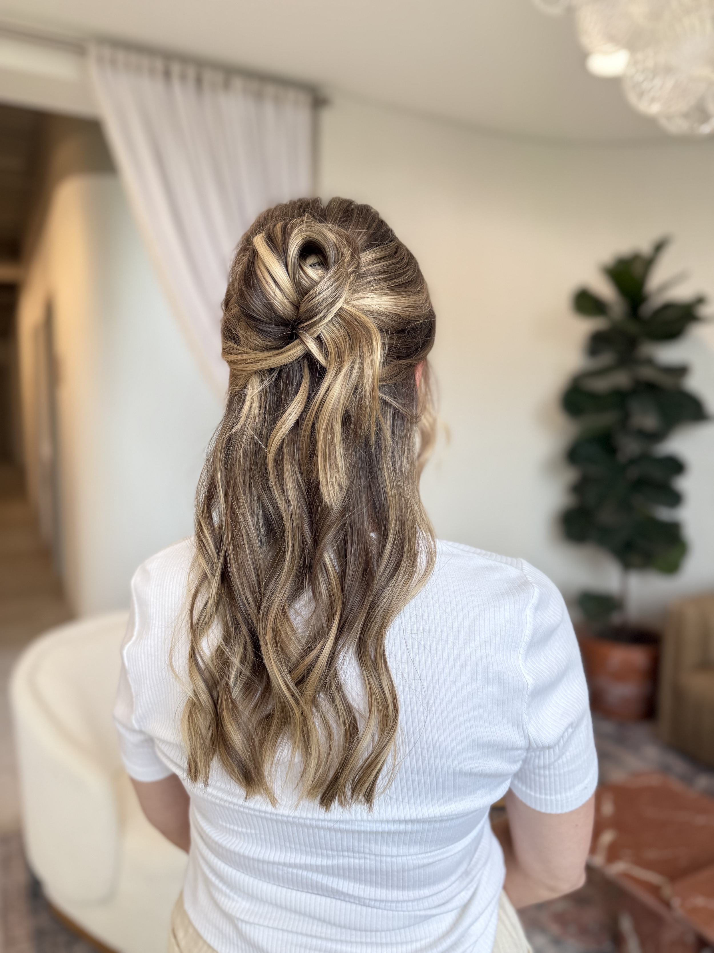 Back view of a woman with long, wavy hair styled in a loose top knot, wearing a white shirt in a room with plants and furniture.
