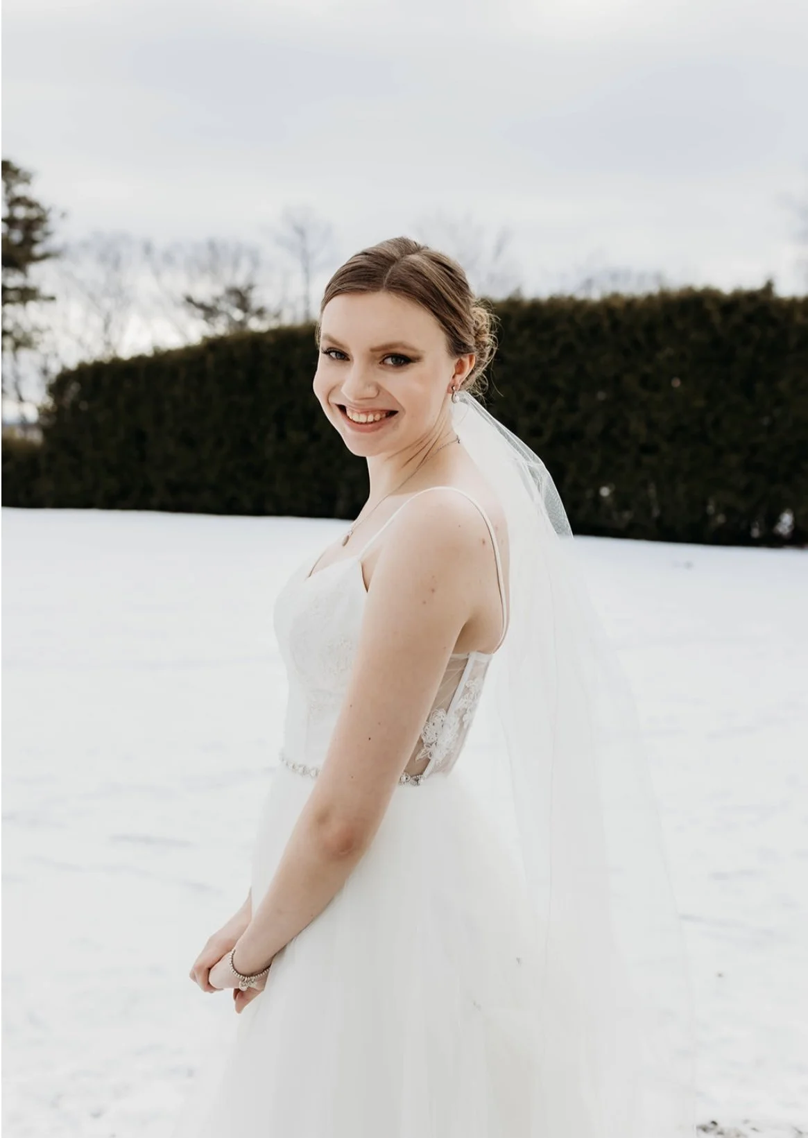 Bride in a white wedding dress smiling outdoors in a snowy landscape with trees and hedges in the background.