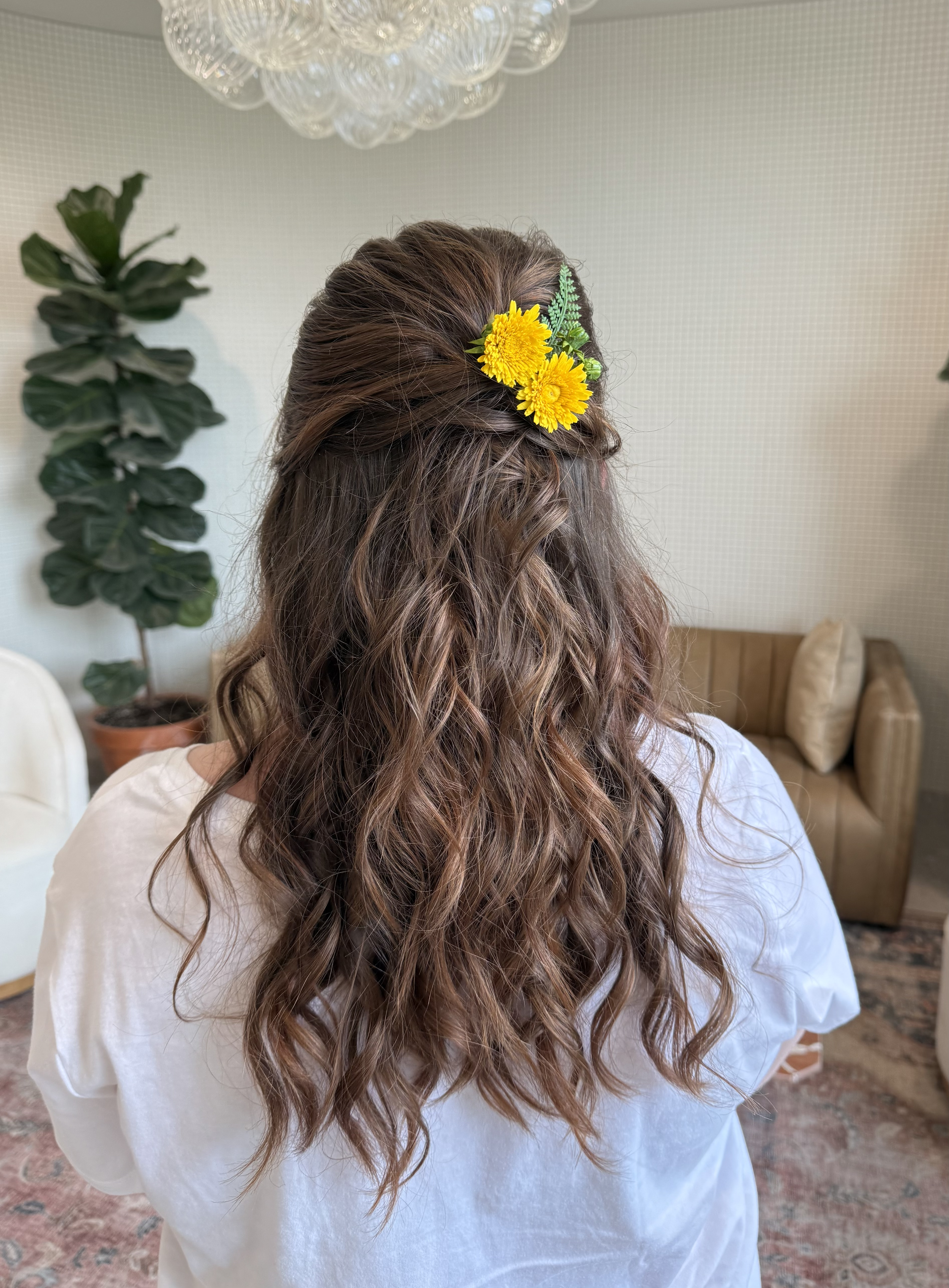 A woman with long, wavy brown hair decorated with a yellow flower hairpiece, seen from behind in a cozy indoor setting with plants and a sofa.