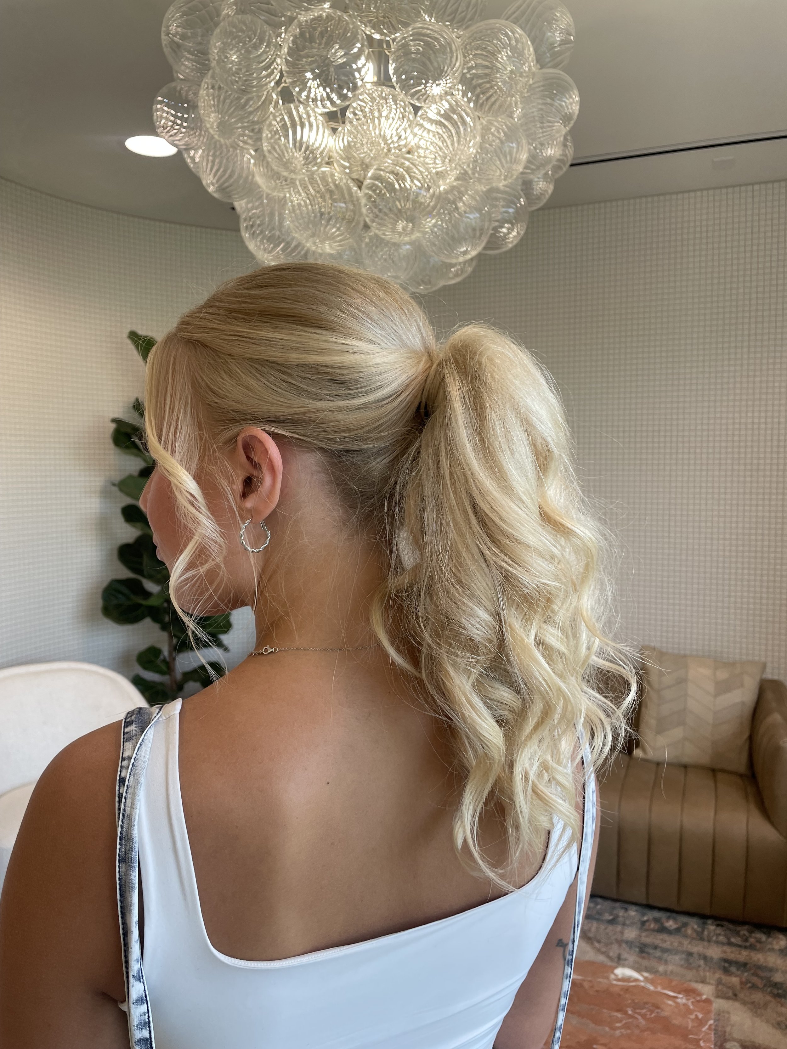 A woman with blonde, curly hair tied in a ponytail, wearing a white tank top and silver jewelry, standing indoors near a modern chandelier made of glass globes, with a background of a white wall, a potted plant, and a brown sofa.