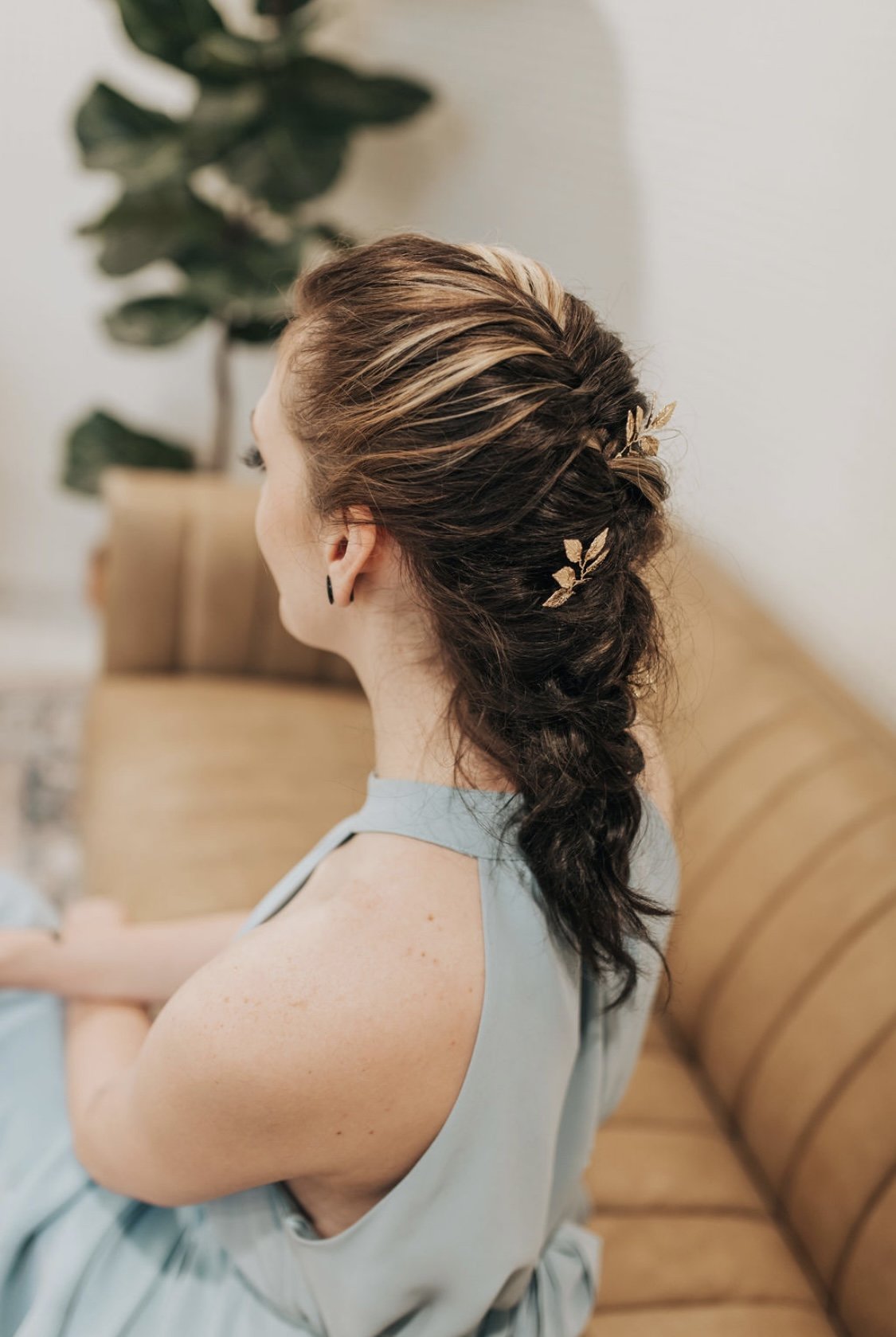 A woman with braided hair and gold leaf hair accessories, sitting on a tan leather couch in a room with a potted plant in the background.