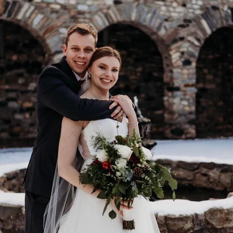 A happy couple in wedding attire posing outdoors near a stone bridge, the man hugging the woman from behind while she holds a bouquet of white flowers and greenery.