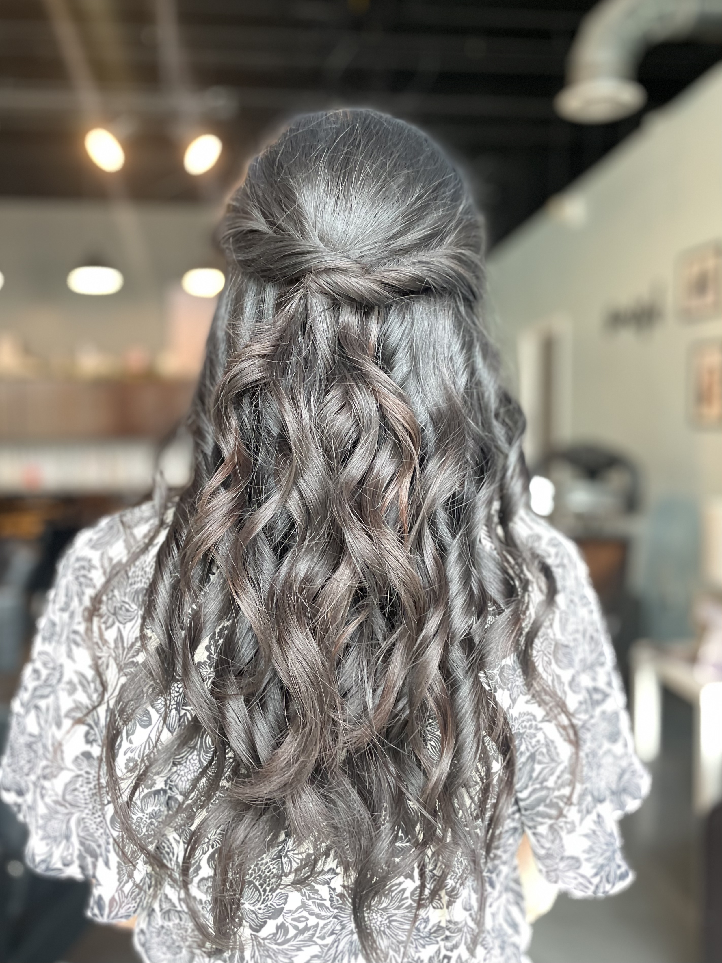 Back view of a woman with long, wavy silver hair styled in a half-up, half-down hairstyle, in an indoor setting.